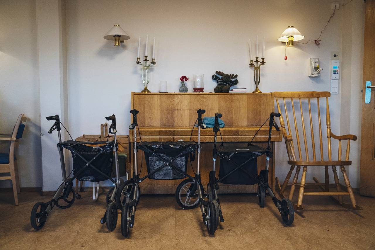 Tre rullatorer står parkert foran et lysebrunt piano. Ved siden av står en gyngestol. På pianoet står to trearmede lysestaker og en elg.