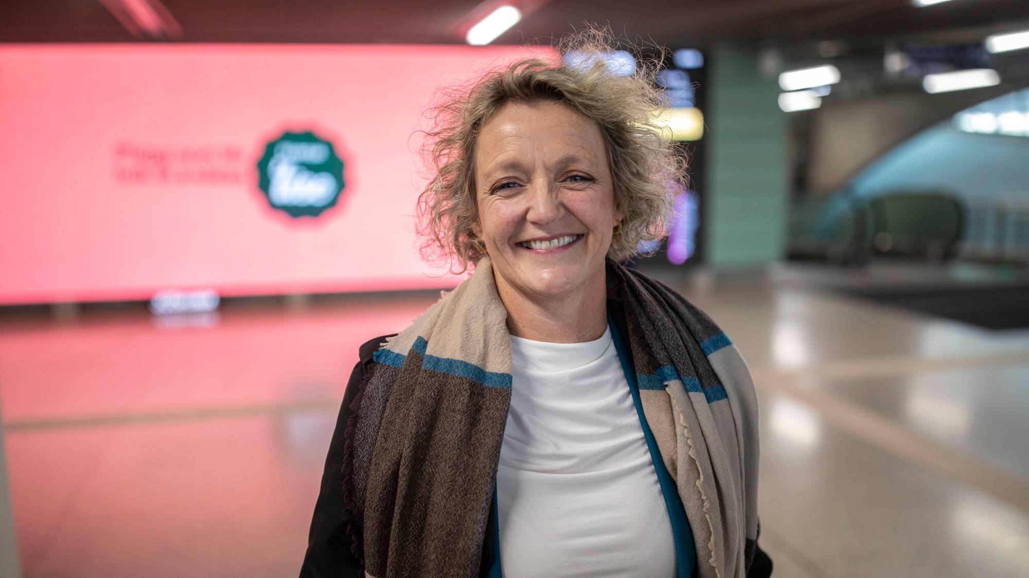 Smilende kvinne med krøllete hår og skjerf, fotografert innendørs i et offentlig rom. (Bildebeskrivelsen er laget av en KI-tjeneste)