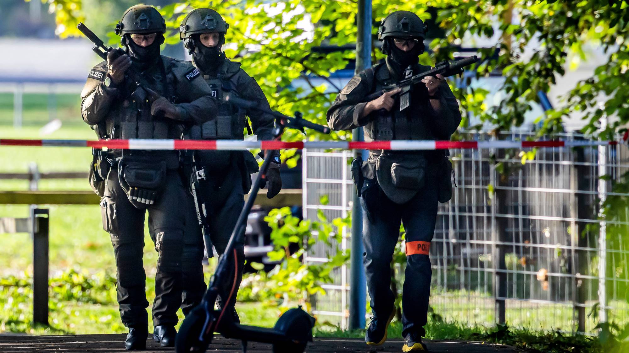 Tre tungt bevæpnede politibetjenter i uniform går bak sperrebånd utendørs. 