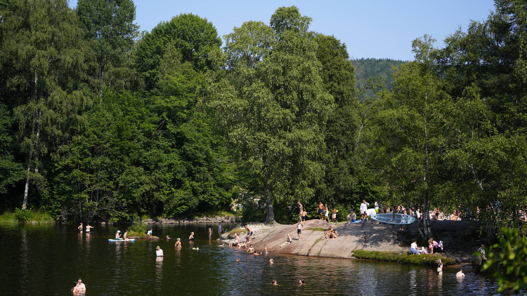 Folk bader og oppholder seg på svaberg ved et skogsvann på en solskinnsdag. (Bildebeskrivelsen er laget av en KI-tjeneste)