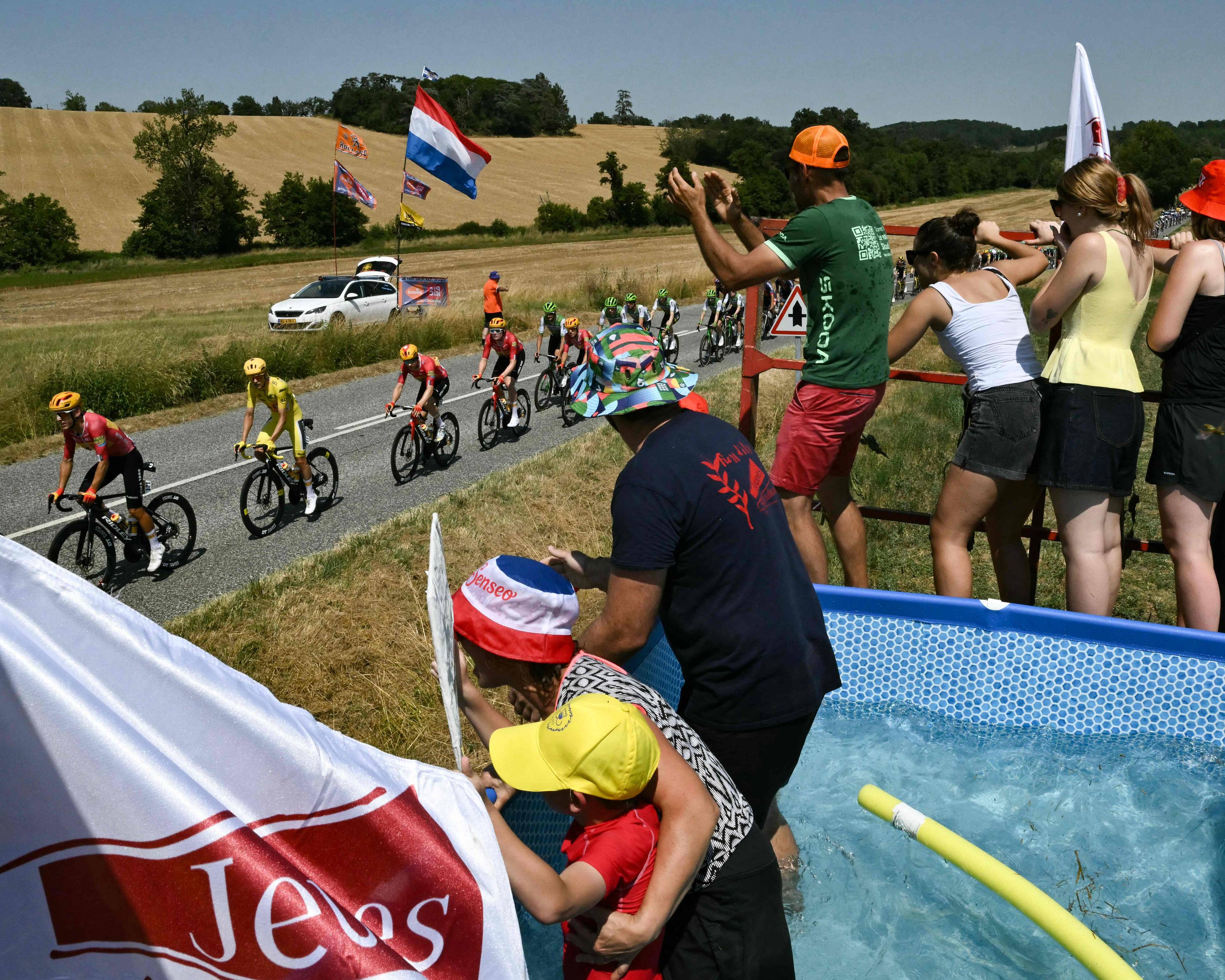 Syklister i Tour de France sykler forbi publikum som står ved et oppblåsbart basseng langs veien. (Bildebeskrivelsen er laget av en KI-tjeneste)