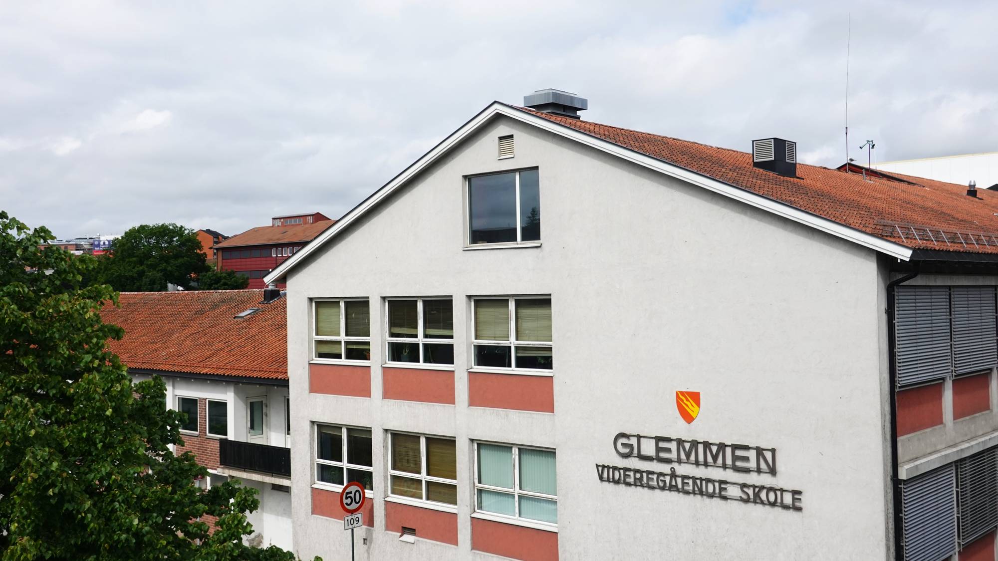Glemmen videregående skole er en bygning med flere vinduer. Et rødt og gult våpenskjold er plassert over tekstskiltet "GLEMMEN VIDEREGÅENDE SKOLE". Et trafikkskilt viser en hastighetsbegrensning på 50. Det er også et tre i forgrunnen. (Bildebeskrivelsen er laget av en KI-tjeneste)