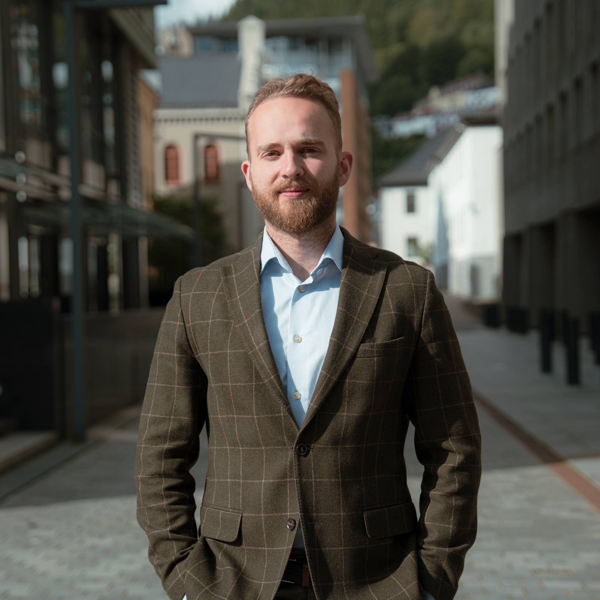 En mann i brun dress og lyseblå skjorte ser muntert inn i kameraet. Han står foran to bygninger. 