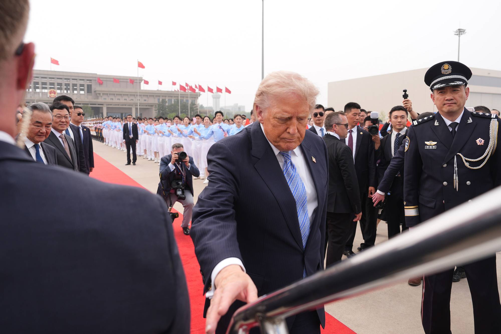 Donald Trump ankommer et arrangement i Kina. Han blir fulgt av medarbeidere og sikkerhetspersonell. En gruppe kvinner i lyseblå uniformer står på rekke og rad i bakgrunnen. (Bildebeskrivelsen er laget av en KI-tjeneste)