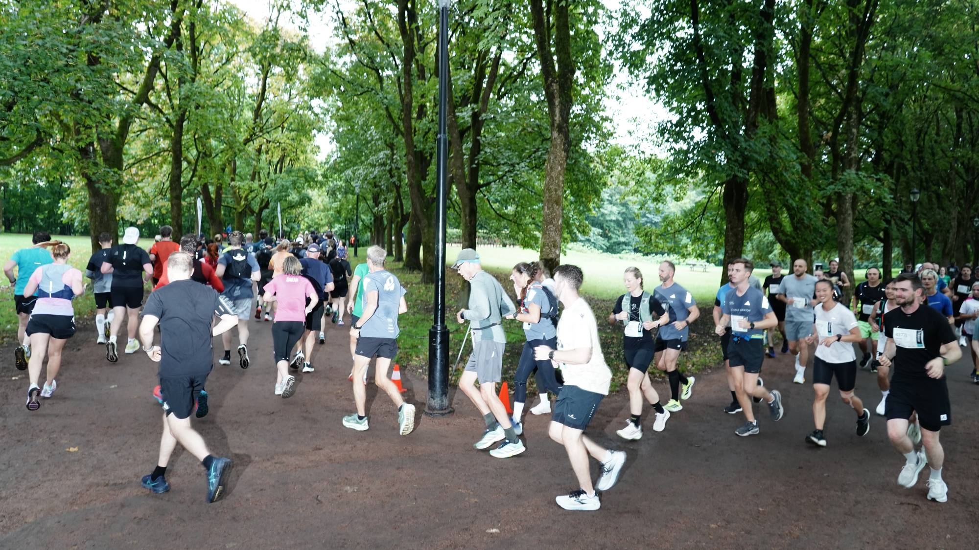 En stor gruppe mennesker løper et mosjonsløp på en grusvei i en park omgitt av trær. (Bildebeskrivelsen er laget av en KI-tjeneste)