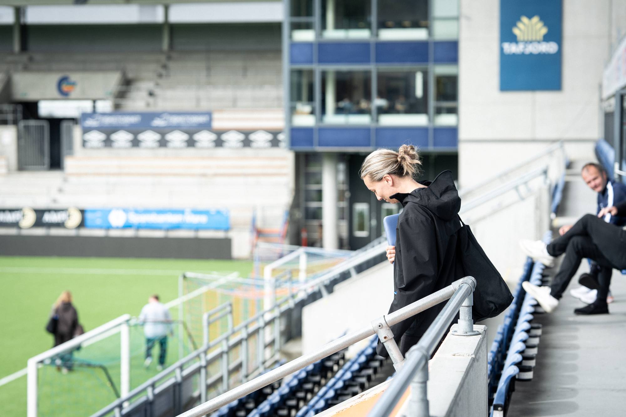 Kvinne med svart jakke går ned trappene på en fotballstadion. Andre personer sitter på tribunen. (Bildebeskrivelsen er laget av en KI-tjeneste)