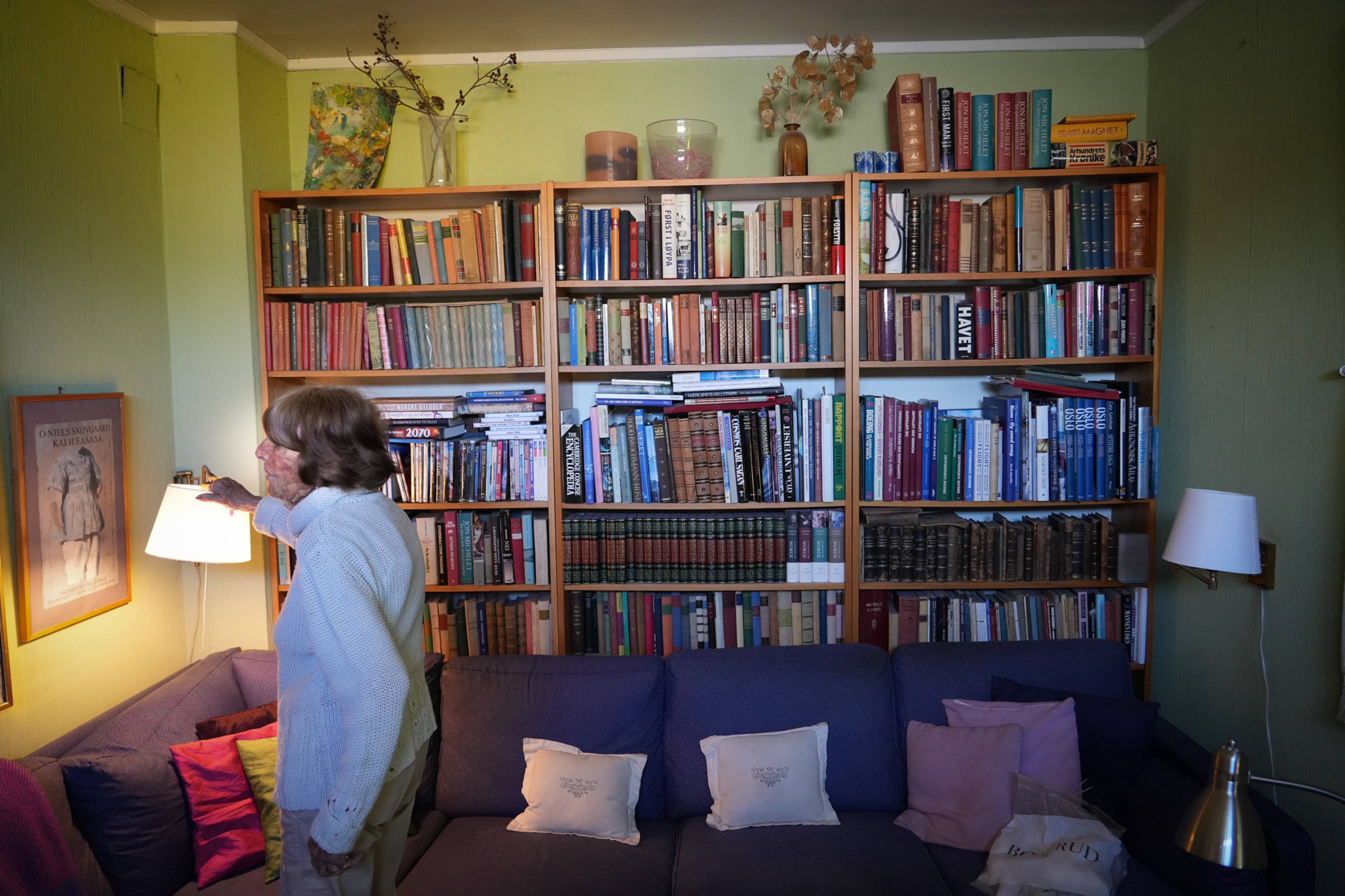En kvinne står foran en bokhylle full av bøker. Hun rekker ut en hånd mot en lampe. Til venstre for henne henger et bilde på veggen. Hun sitter i en sofa. (Bildebeskrivelsen er laget av en KI-tjeneste)