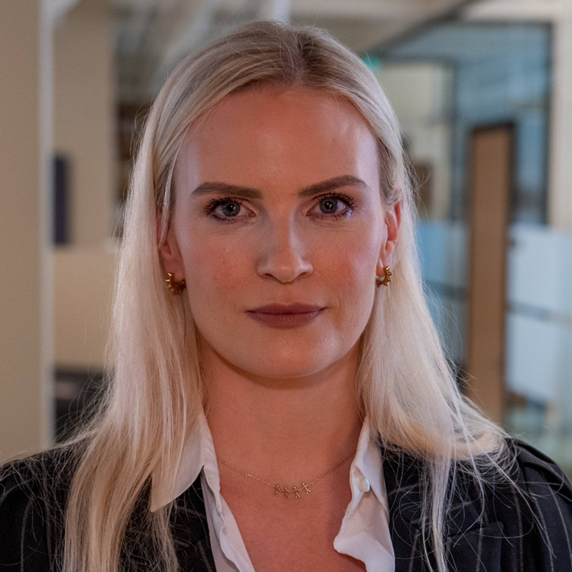 Kvinne med blondt hår i mørk dressjakke står innendørs i et kontorlandskap. 