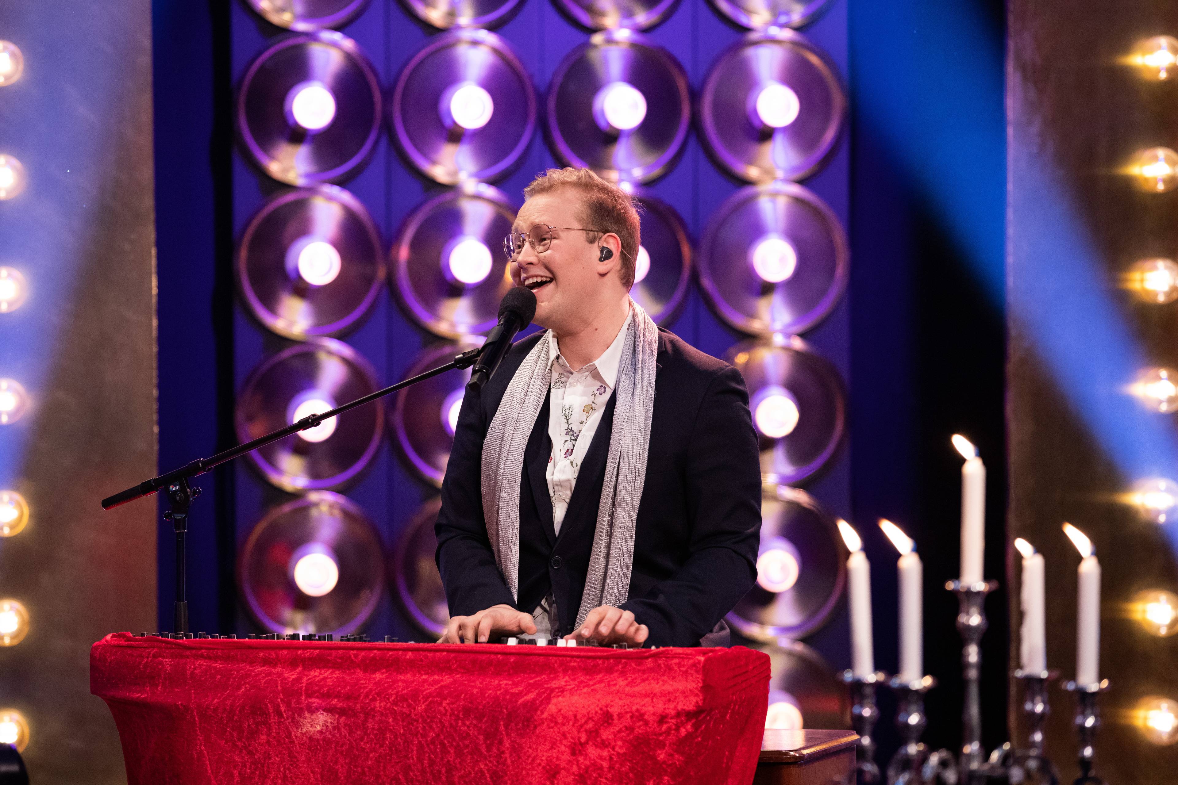Mann med dress og glitrande skjerf sit og syng i ein mikrofon med eit piano trekt med raud fløyelsduk.