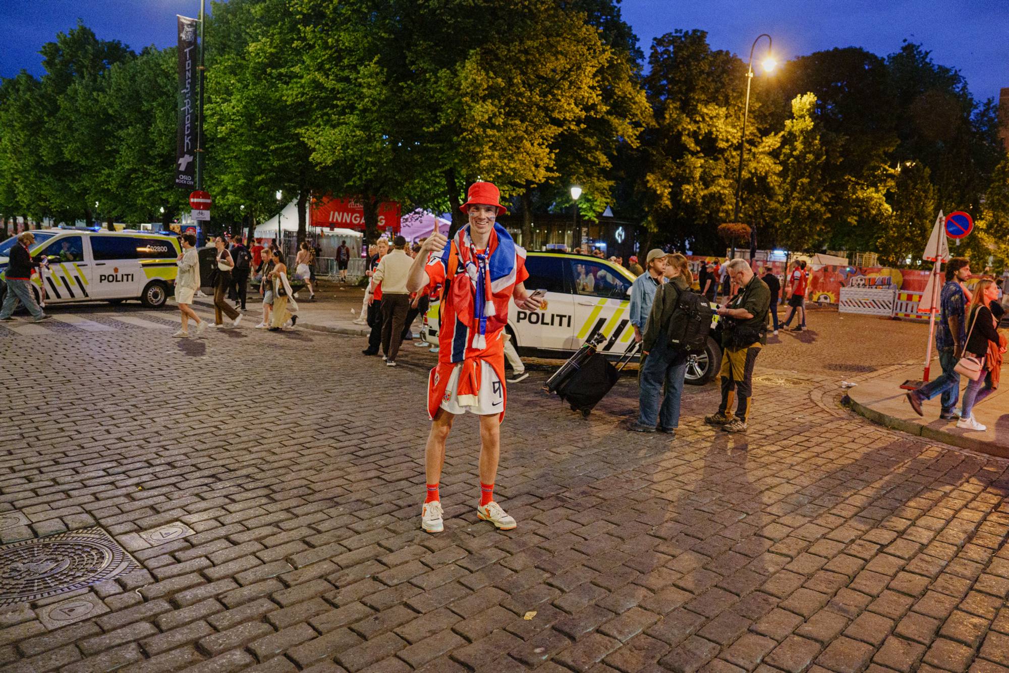 Supporter i norske farger gir tommel opp ved to politibiler på en bygate. (Bildebeskrivelsen er laget av en KI-tjeneste)