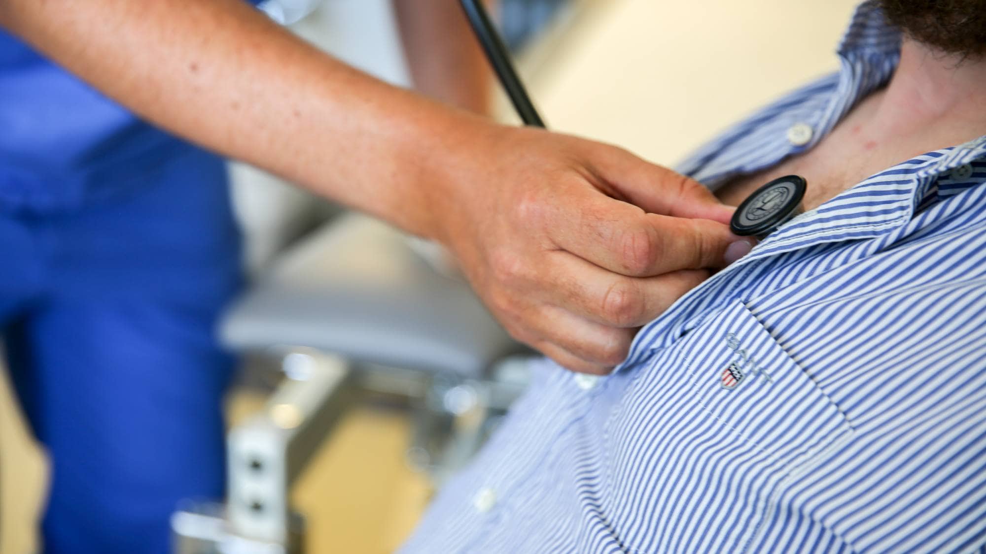 Ein person med skjegg sit i ein stol med to-tre knappar kneppa opp. Ein lege held eit stetoskop til brystet. Vi ser korkje av dei sitt andlet. I bakgrunnen, ute av fokus, ser vi stolar og utstyr som høyrer til på eit legekontor.