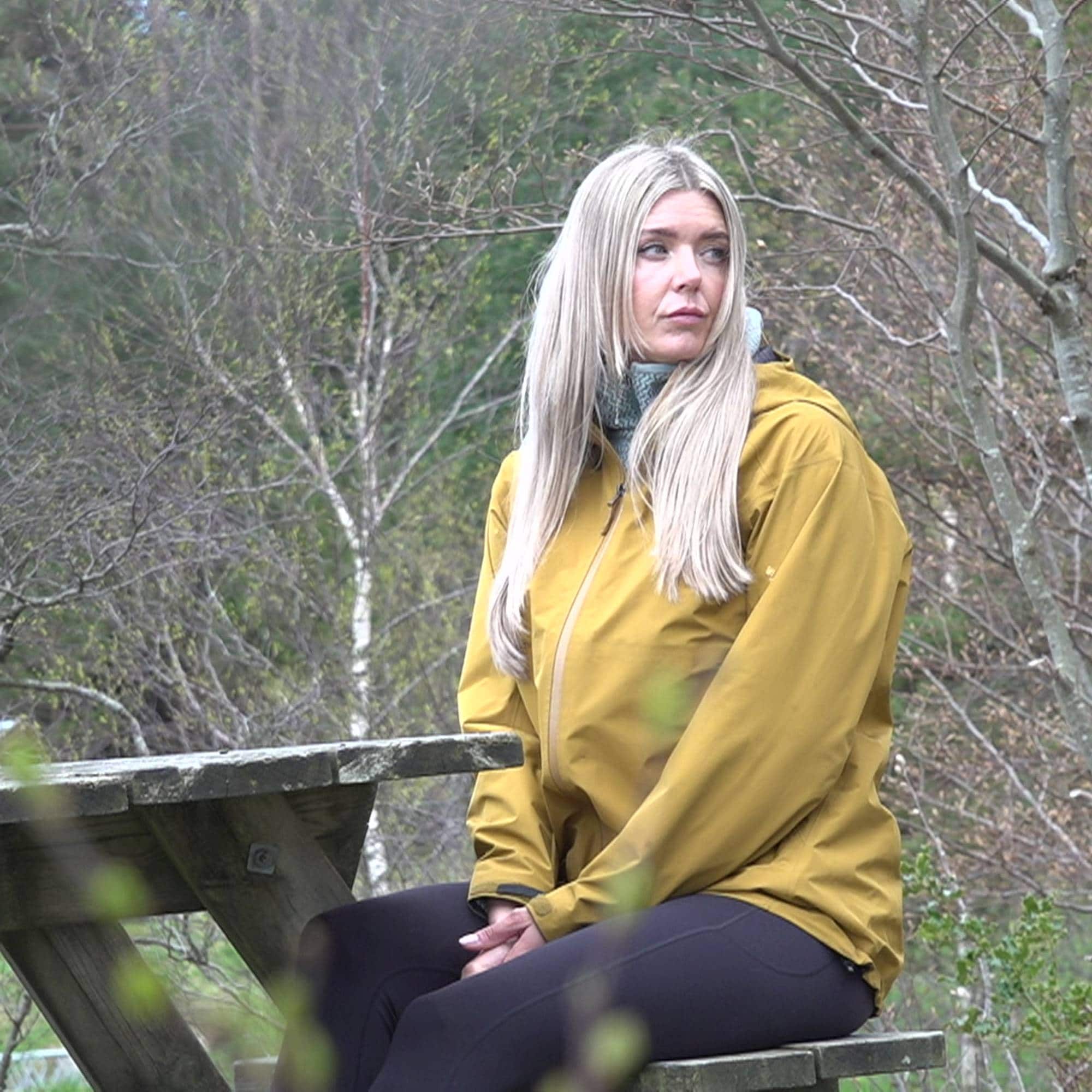 En kvinne sitter på en benk utendørs. Hun har på seg en gul jakke og mørke treningsbukser. Kvinnen har blondt hår og ser til siden. Bak henne er det trær og vegetasjon. (Bildebeskrivelsen er laget av en KI-tjeneste)
