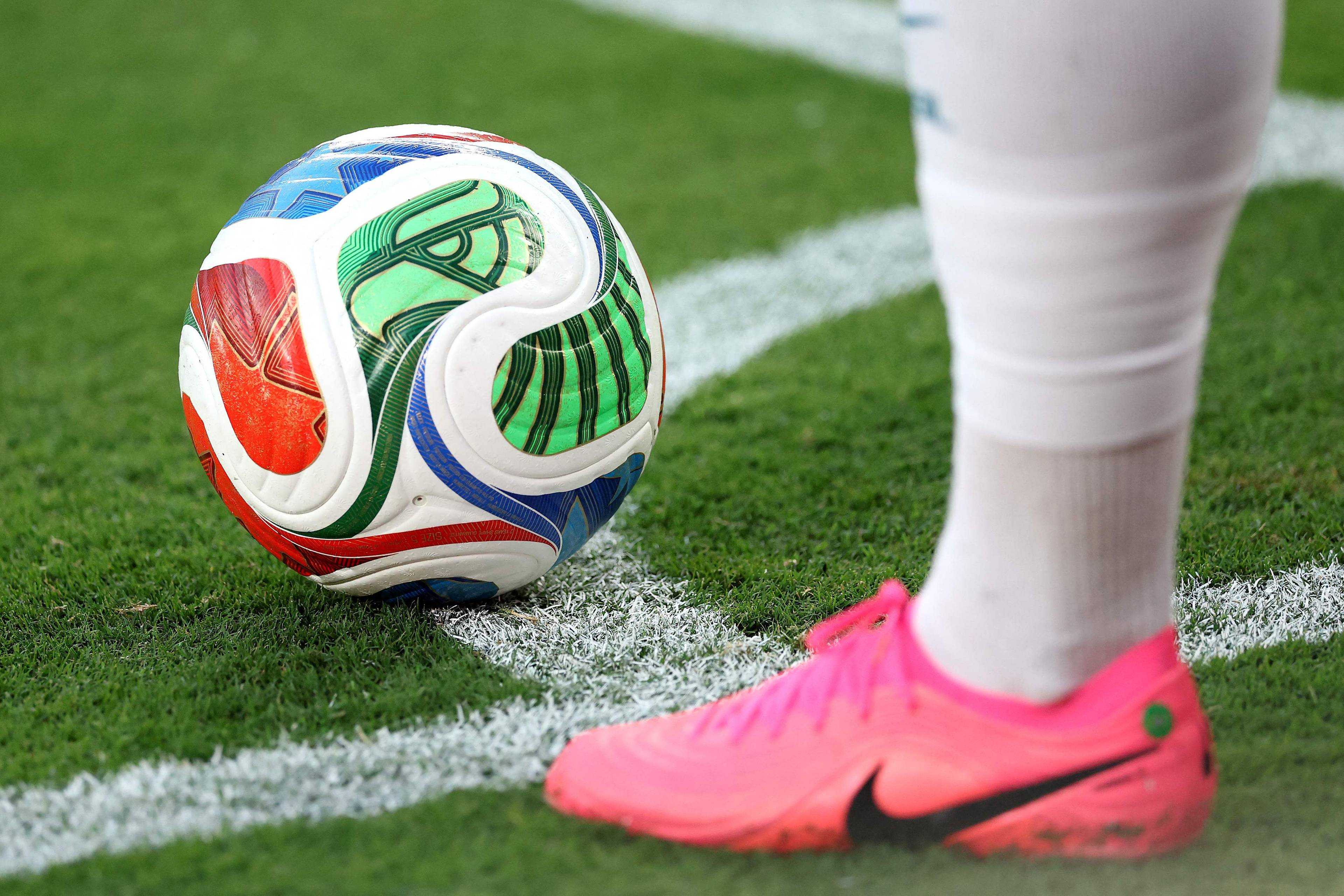 Nærbilde av en fotball på gressmatte med hvit markering. En person i hvite sokker og rosa sko står ved siden av ballen. Et vannmerke er synlig over ballen. (Bildebeskrivelsen er laget av en KI-tjeneste)