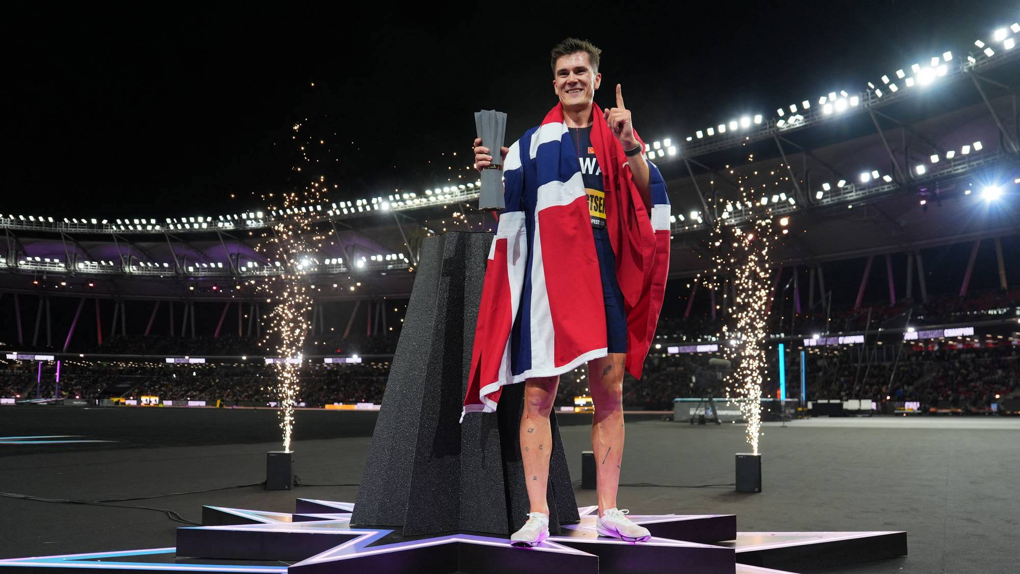 Jakob Ingebrigtsen feirer gullmedalje på en stjerneformet podie med det norske flagget og pokal. (Bildebeskrivelsen er laget av en KI-tjeneste)
