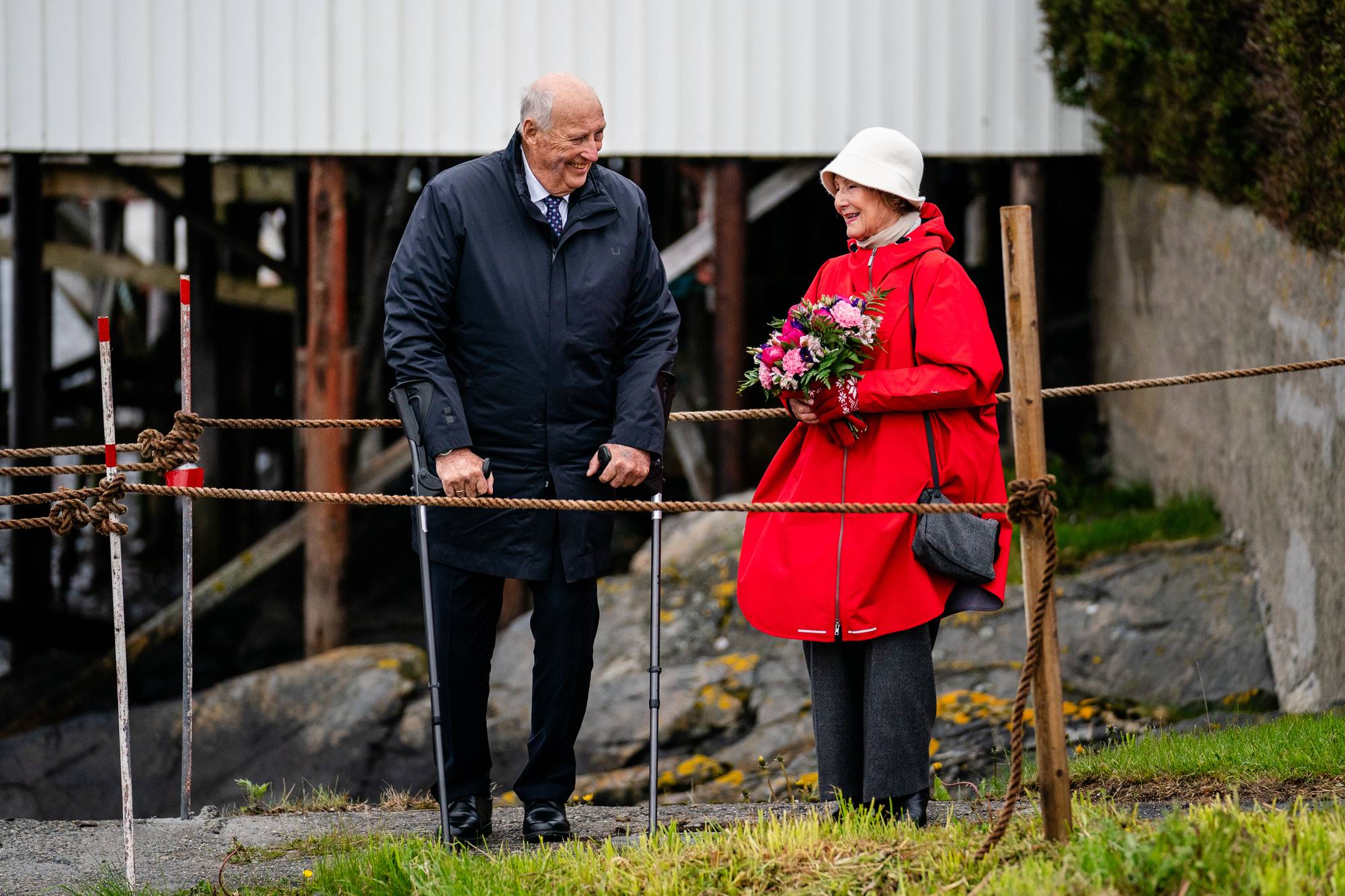Kong Harald ser på dronning Sonja og smiler under besøket i Sømna på fylkesreisen i 2023. Dronningen er ikledd en rød regnjakke og hvit hatt, mens kongen har en blå frakk og lener seg mot krykkene.