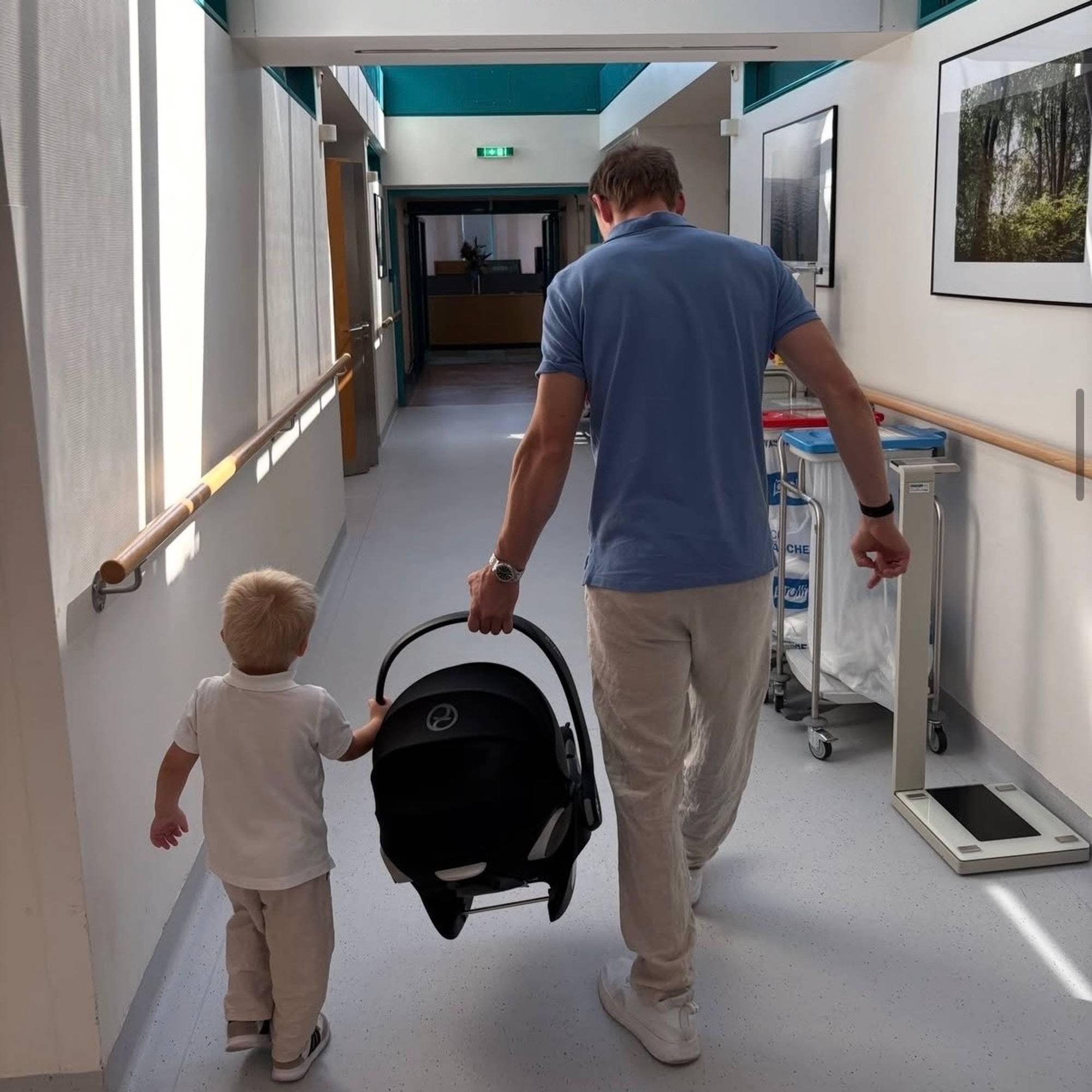 En mann og et barn går bortover en sykehuskorridor. Mannen bærer på en bilstol for baby. (Bildebeskrivelsen er laget av en KI-tjeneste)
