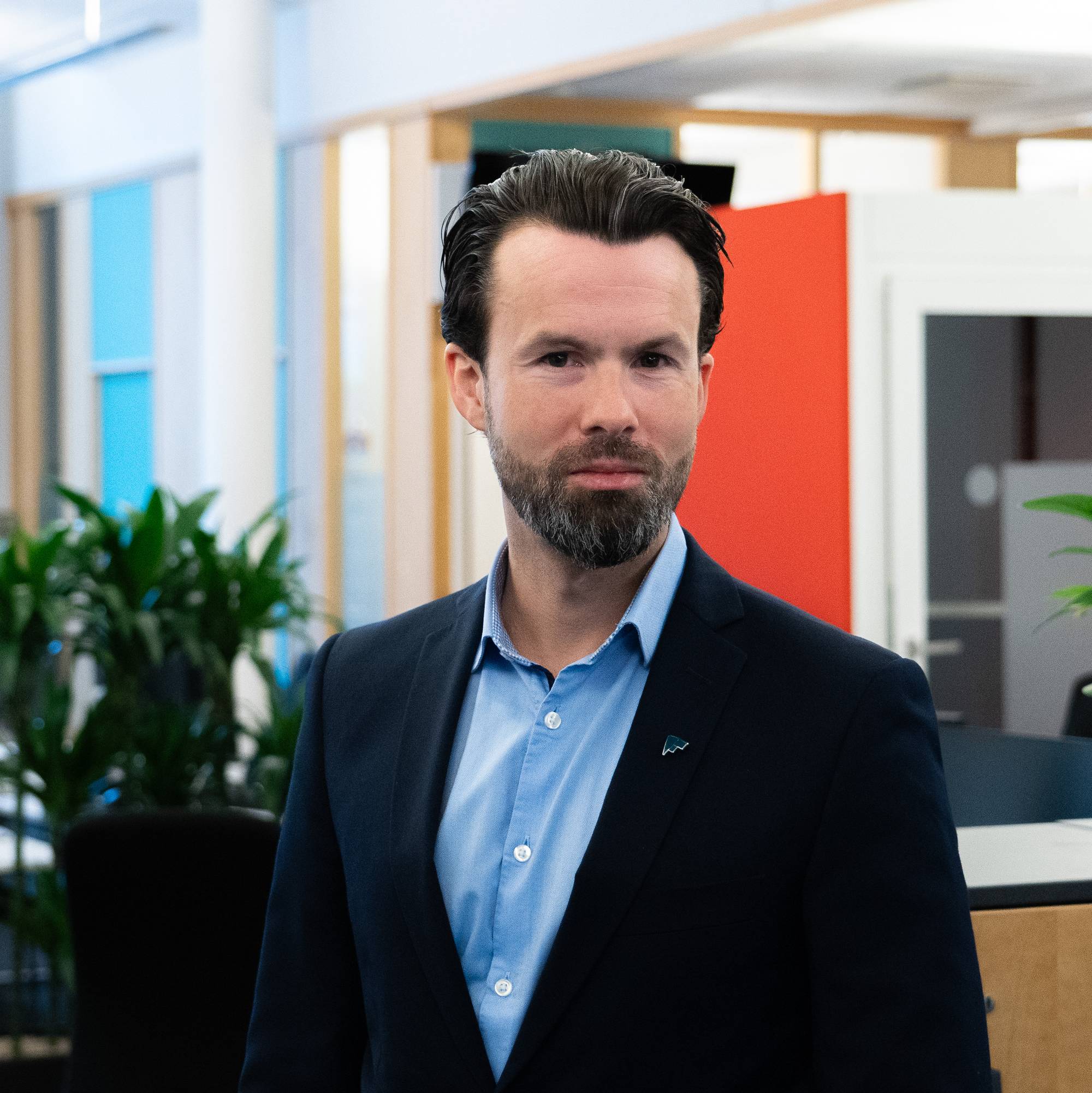 En mann i mørk dress og lyseblå skjorte er fotografert innenfra et kontorlokale. Mannen har mørkt hår og skjegg. Bak mannen er det planter og kontorinnredning. (Bildebeskrivelsen er laget av en KI-tjeneste)