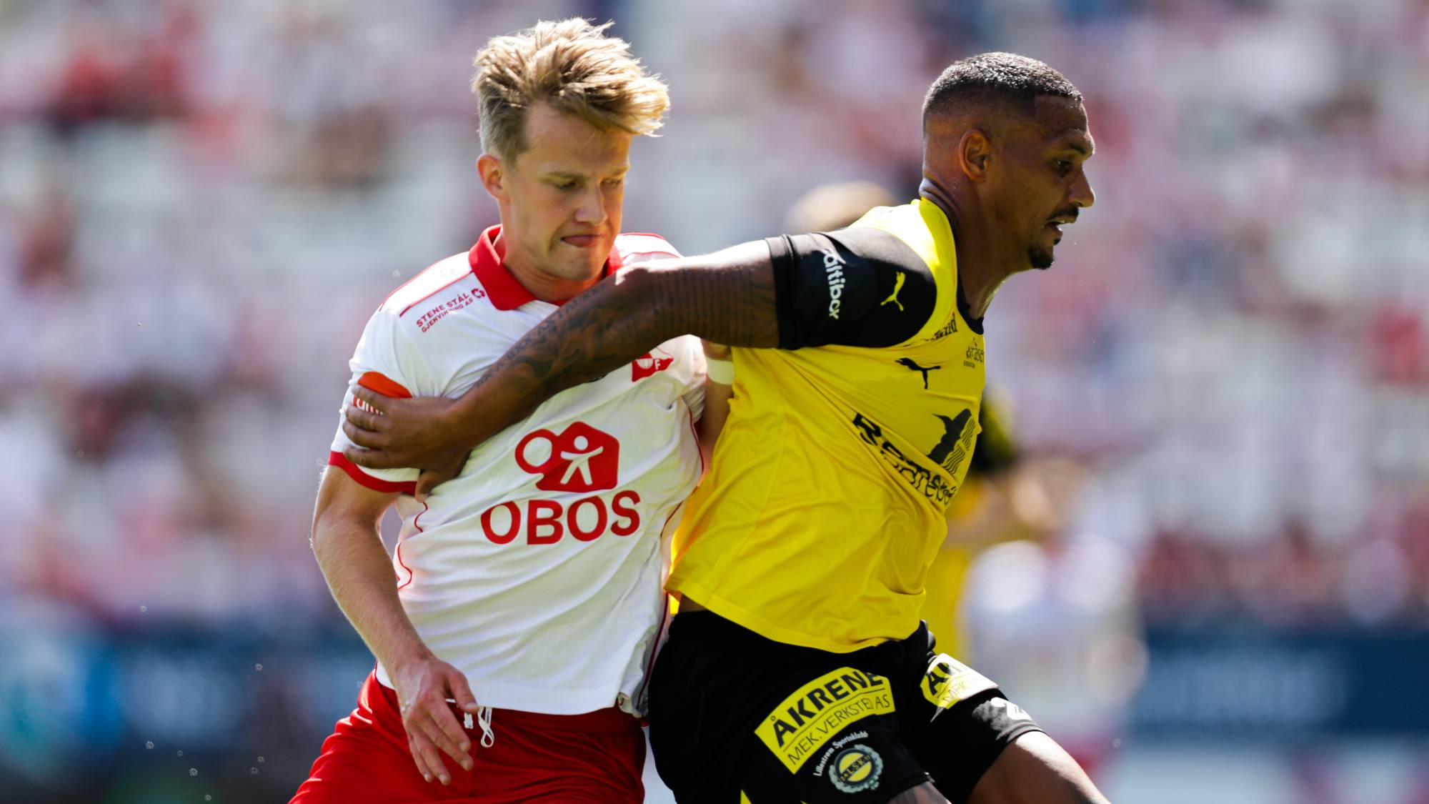 To fotballspillere i duell under en kamp på en stadion. (Bildebeskrivelsen er laget av en KI-tjeneste)