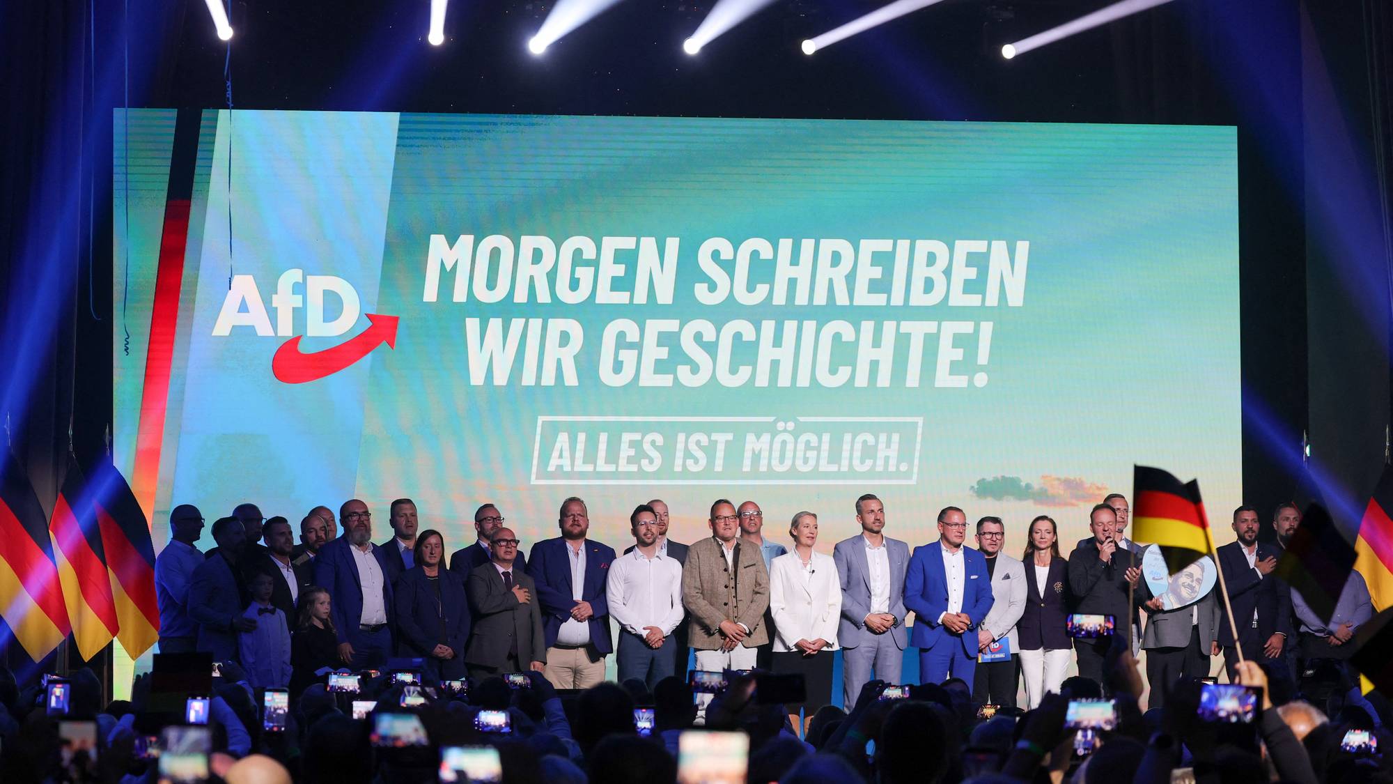 Politikere fra partiet AfD står på en scene foran en storskjerm med teksten Morgen schreiben wir Geschichte. (Bildebeskrivelsen er laget av en KI-tjeneste)
