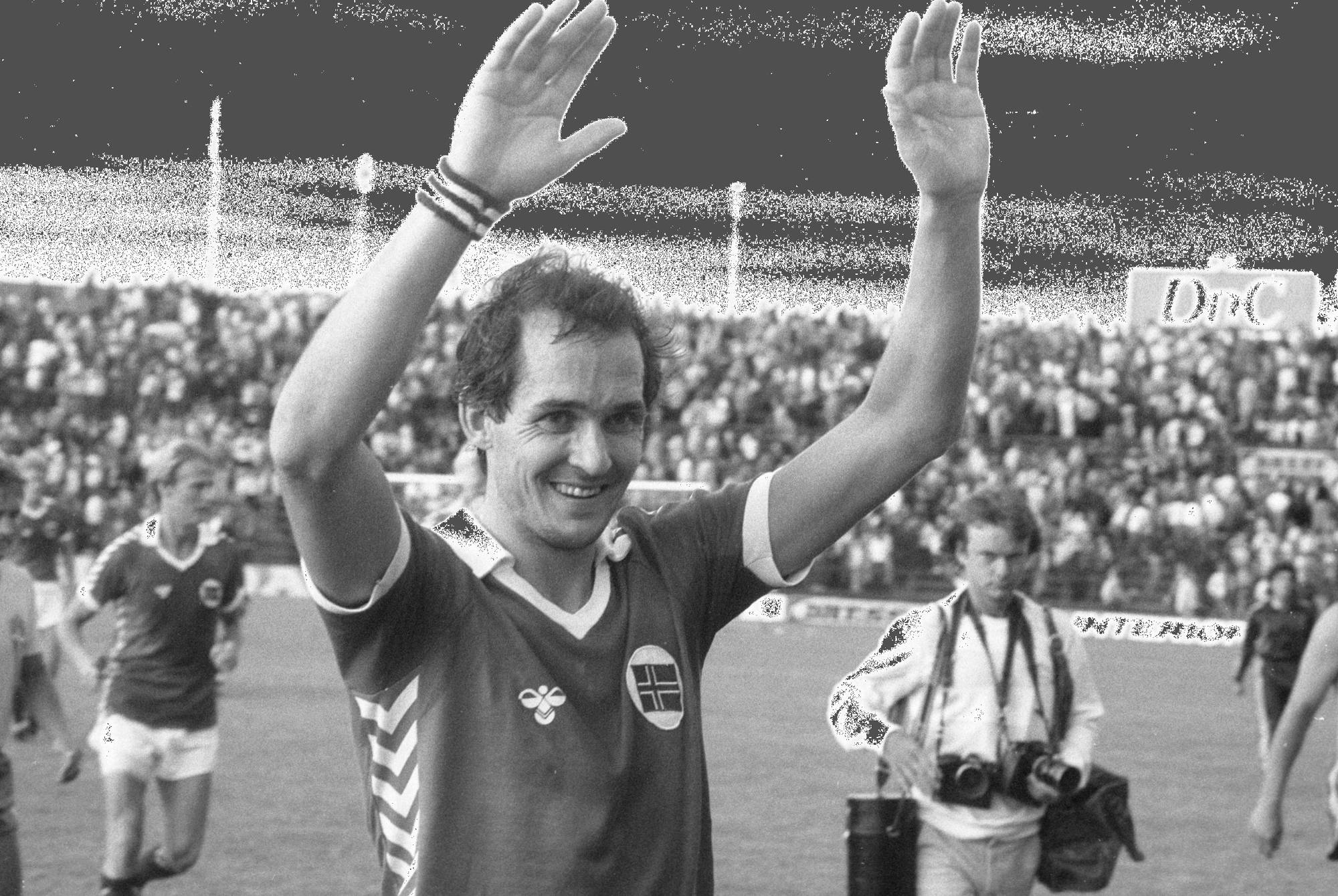 En fotballspiller vinker til publikum. Han er iført en rød trøye med et norsk flagg på brystet. I bakgrunnen står tilskuere på tribunen. En annen person med kamera går forbi. (Bildebeskrivelsen er laget av en KI-tjeneste)