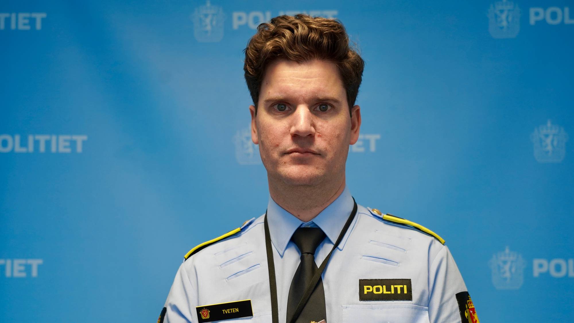 En mann i politiuniform foran en blå vegg med Politiets logo.
