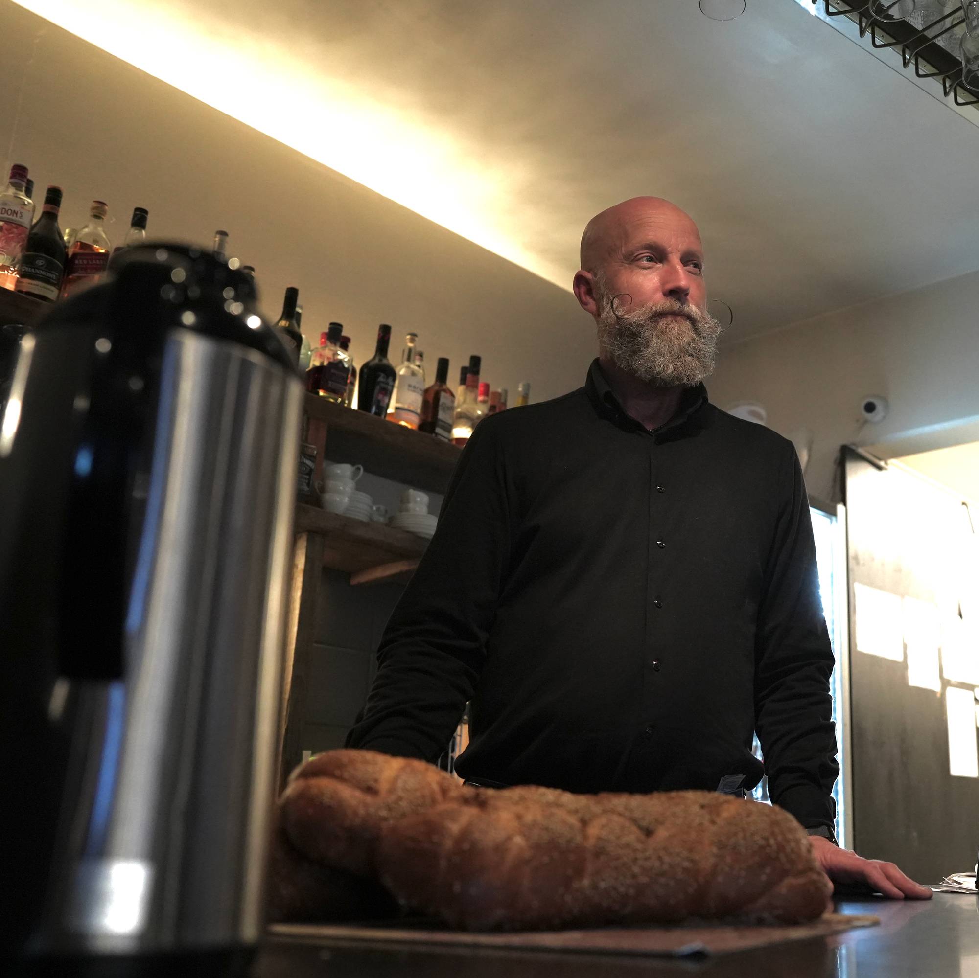 En mann står bak en disk i en restaurant. Han er kledd i en svart skjorte. Mannen er skallet med grå skjegg.