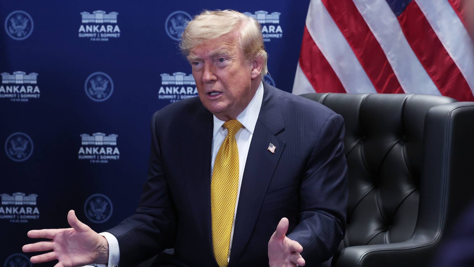 Donald Trump sitter i en stol og snakker foran et amerikansk flagg og en bakgrunn med logoer. 