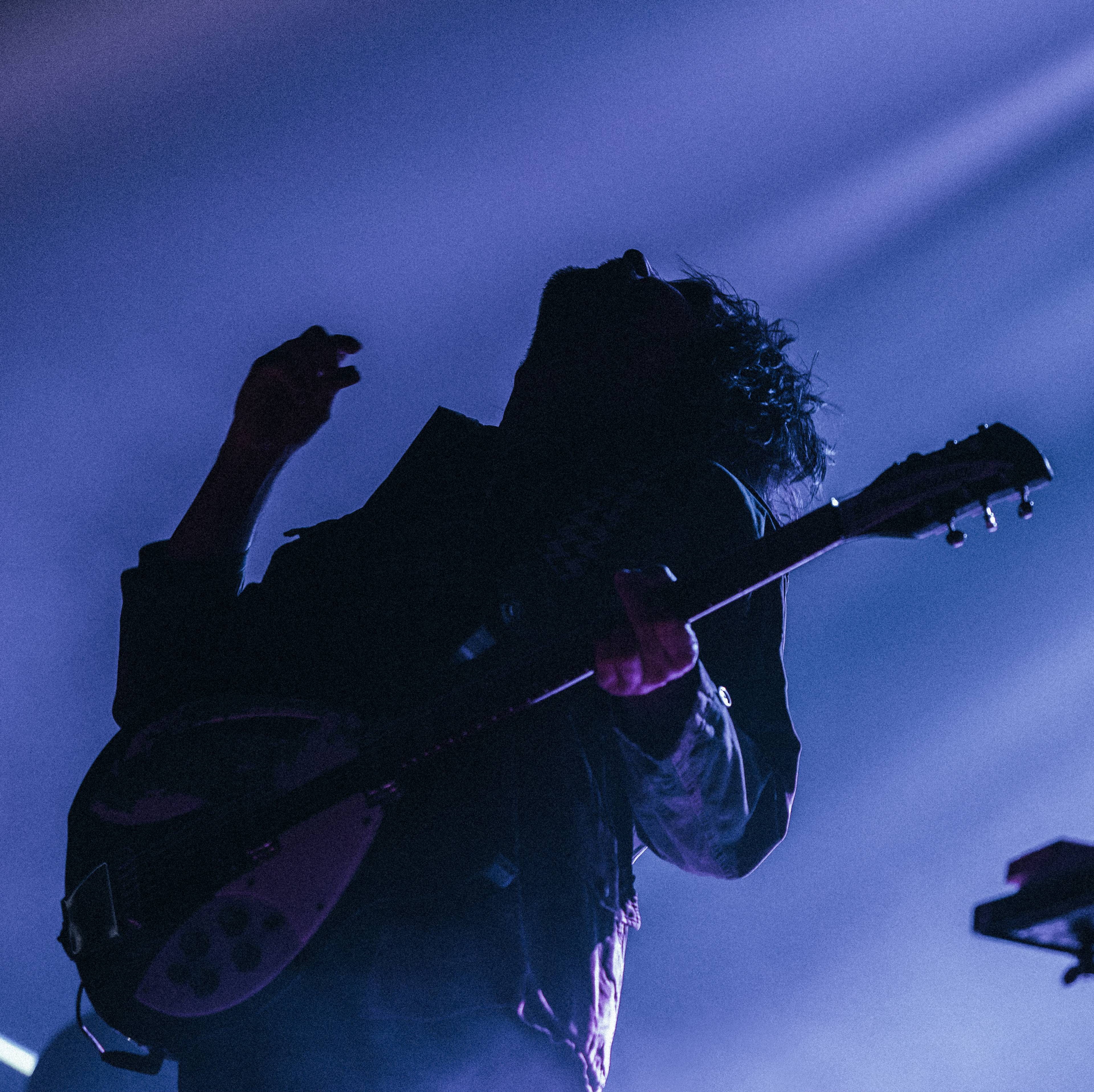 En mann  i motlys spiller gitar med innlevelse og ser opp mot scenelyset over