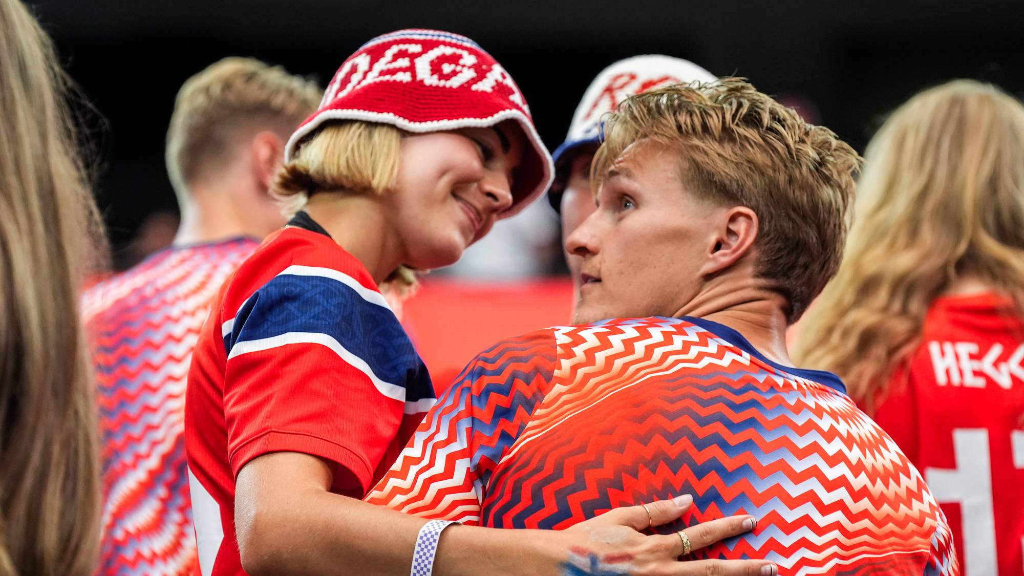 Supporter i rød hatt holder rundt fotballspiller Martin Ødegaard på en tribune. (Bildebeskrivelsen er laget av en KI-tjeneste)