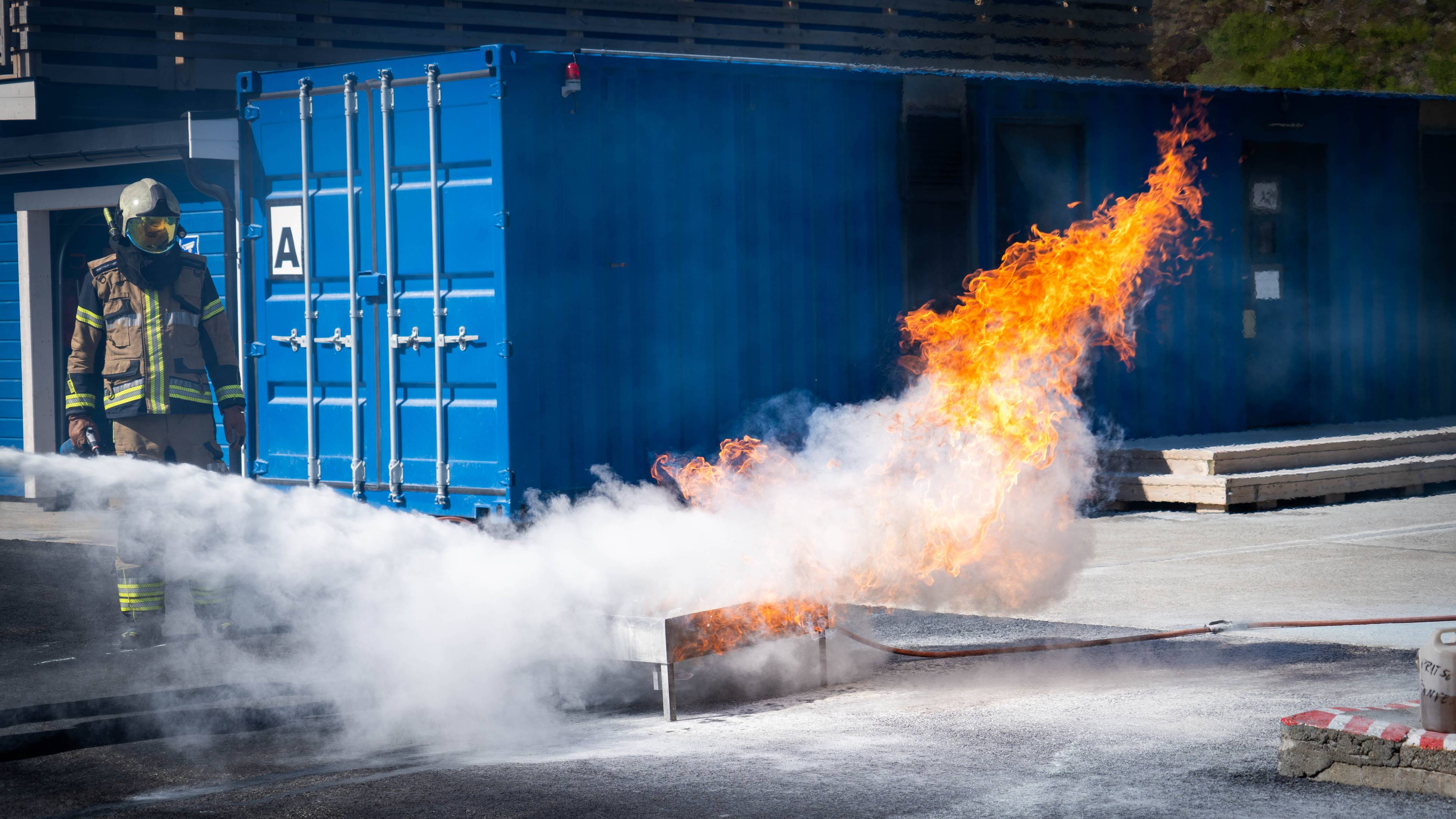 Brannkonstabel i fullt verneutstyr slukker en flamme foran en blå container. (Bildebeskrivelsen er laget av en KI-tjeneste)