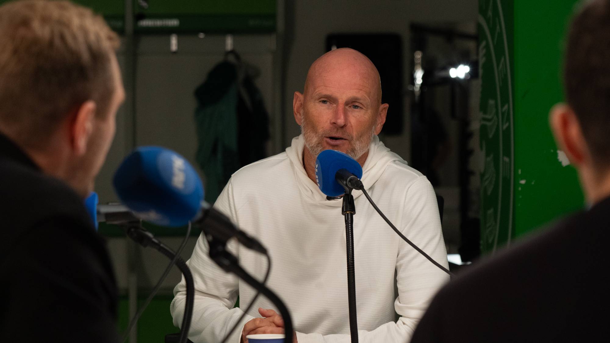 Ståle Solbakken snakker i mikrofon under et intervju med to personer i forgrunnen. (Bildebeskrivelsen er laget av en KI-tjeneste)