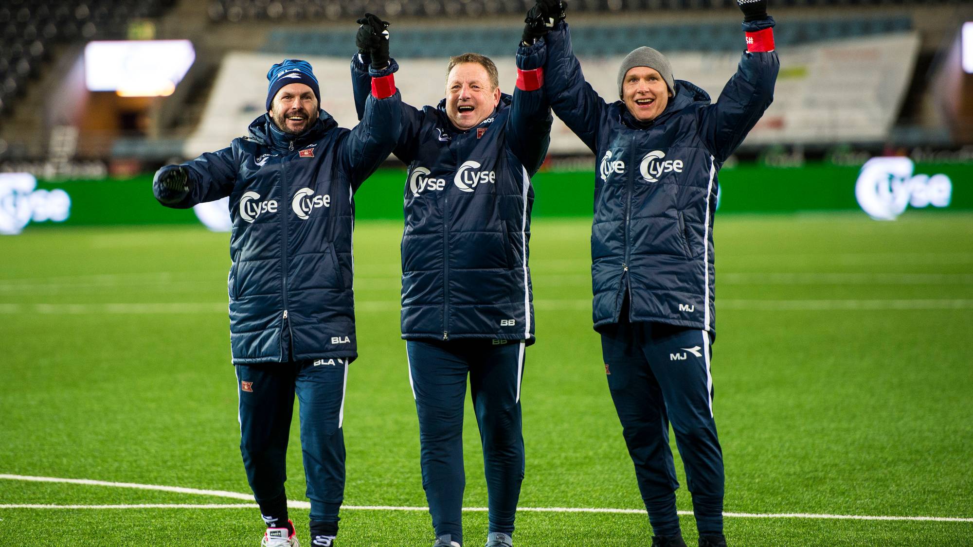 Tre personer i treningstøy står på en fotballbane og hever hendene i triumf. De smiler og viser glede. Bakgrunnen viser en stadion med tomme seter. Det er belysning og kunstgress på banen.