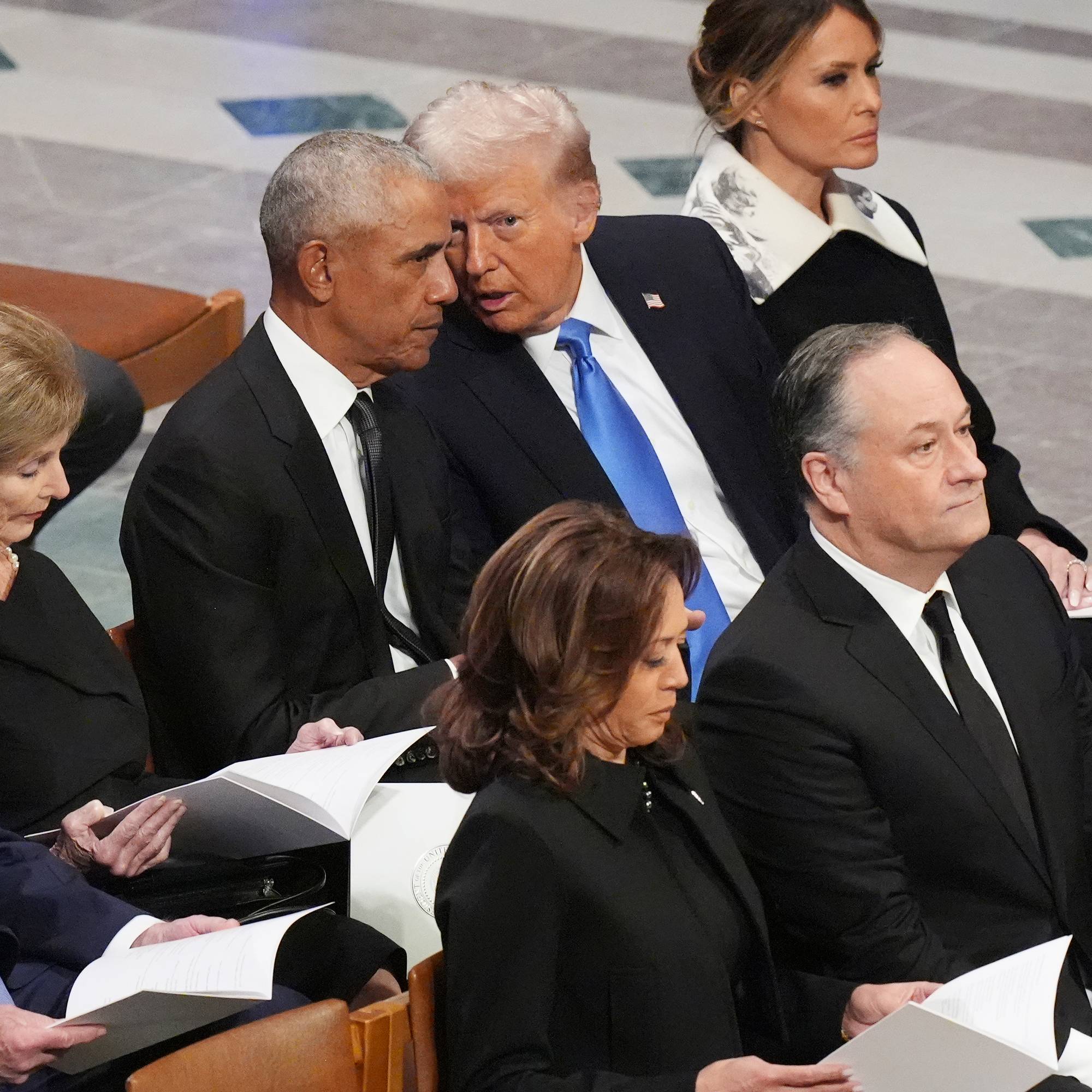 Donald Trump lener seg til siden for å si noe til Barack Obama. Rundt dem sitter Melania Trump, George W. Bush, Laura Bush, Kamala Harris. De er i en begravelse.