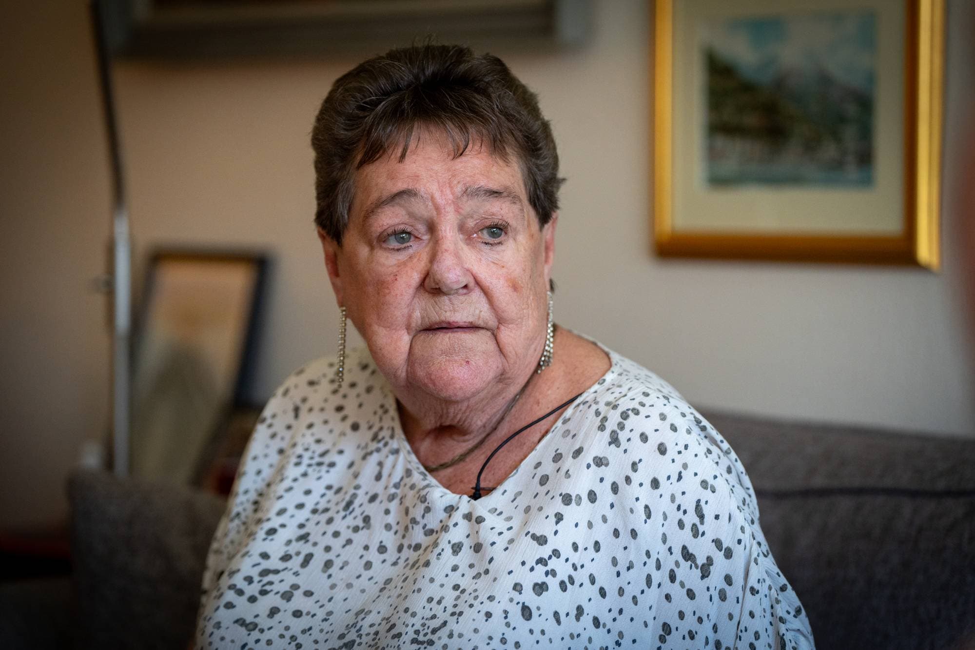 En kvinne med mørkt hår og hvit topp kikker ut av vinduet. I bakgrunnen ser man et bilde.