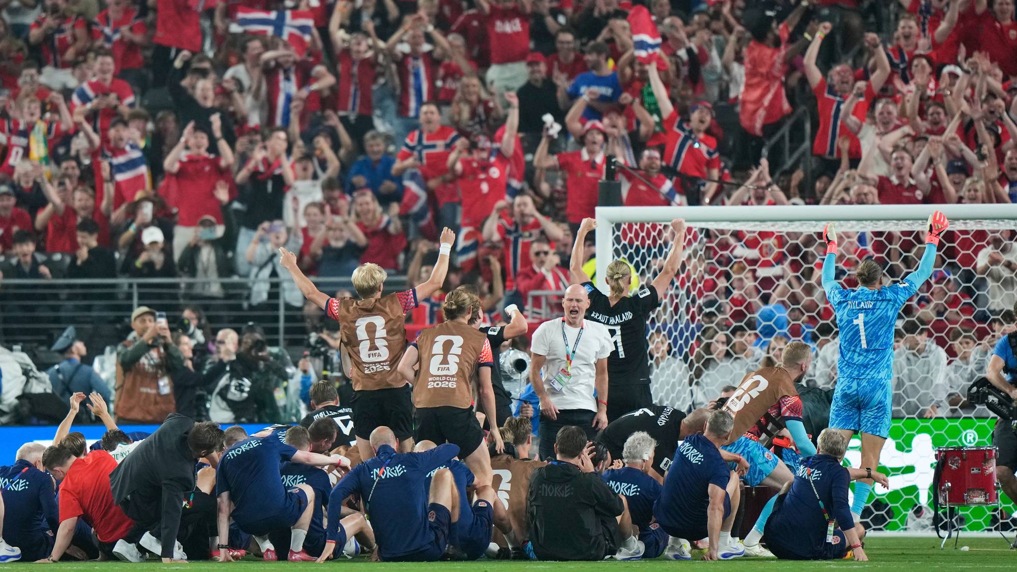 Supportere med norsk flagg jubler på tribunen bak et fotballag. Flere spillere med nummer på ryggen sitter på bakken foran et mål. En keeper i blå drakt har armene i været. (Bildebeskrivelsen er laget av en KI-tjeneste)
