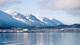 Benjamin Fredriksen Bilde av en kystlinje med snødekte fjell i bakgrunnen. I forgrunnen er det et stille hav som reflekterer omgivelsene. Det finnes flere bygninger i ulike farger på land, samt små båter i havnen. Himlen er blå med noen lette skyer. (Bildebeskrivelsen er laget av en KI-tjeneste)