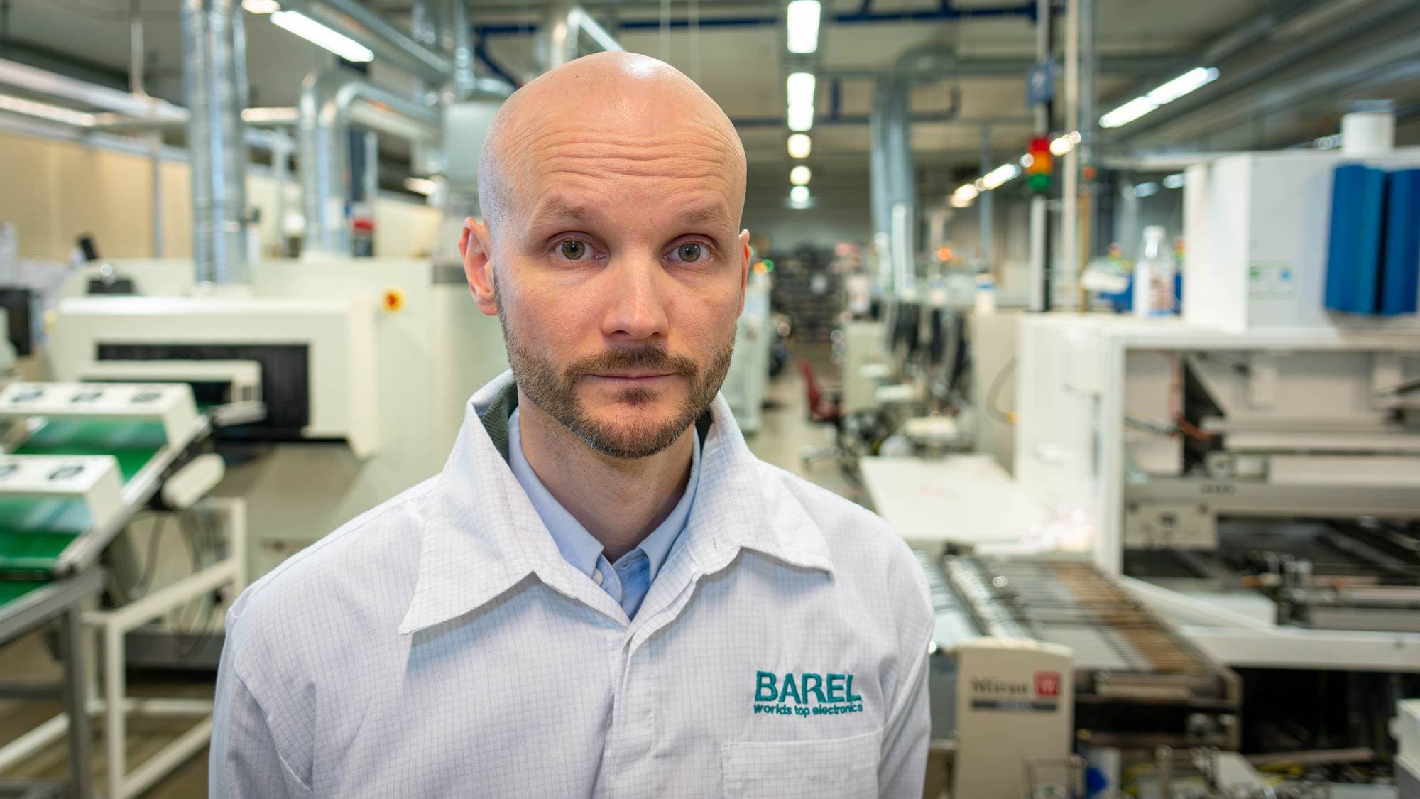 Skallet mann med skjegg i hvit arbeidskjorte står foran produksjonsmaskiner i en fabrikk. (Bildebeskrivelsen er laget av en KI-tjeneste)