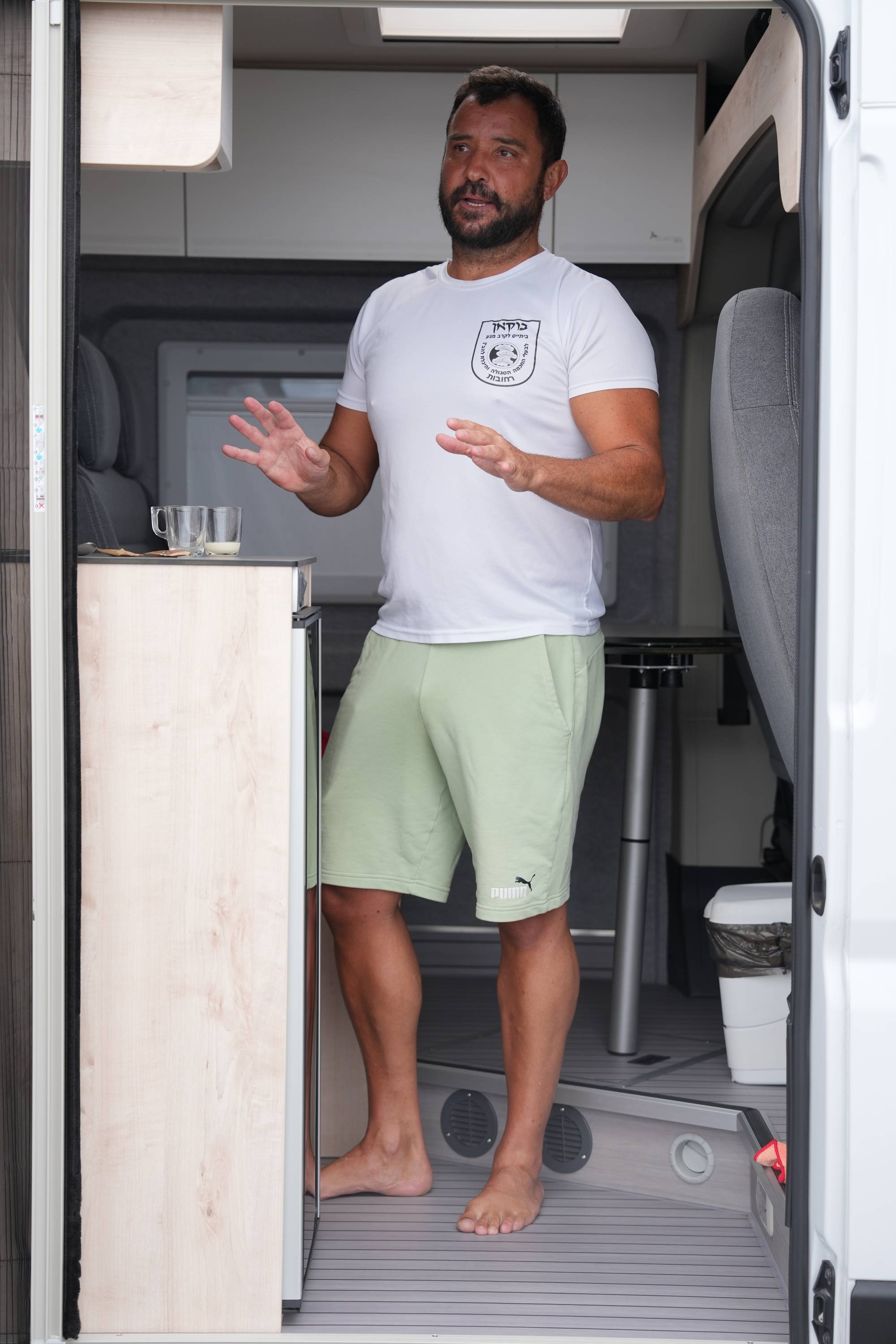 En mann står i døråpningen til en bobil. Han har på seg en hvit t-skjorte og lysegrønn shorts. Mannen har hendene løftet og snakker. 