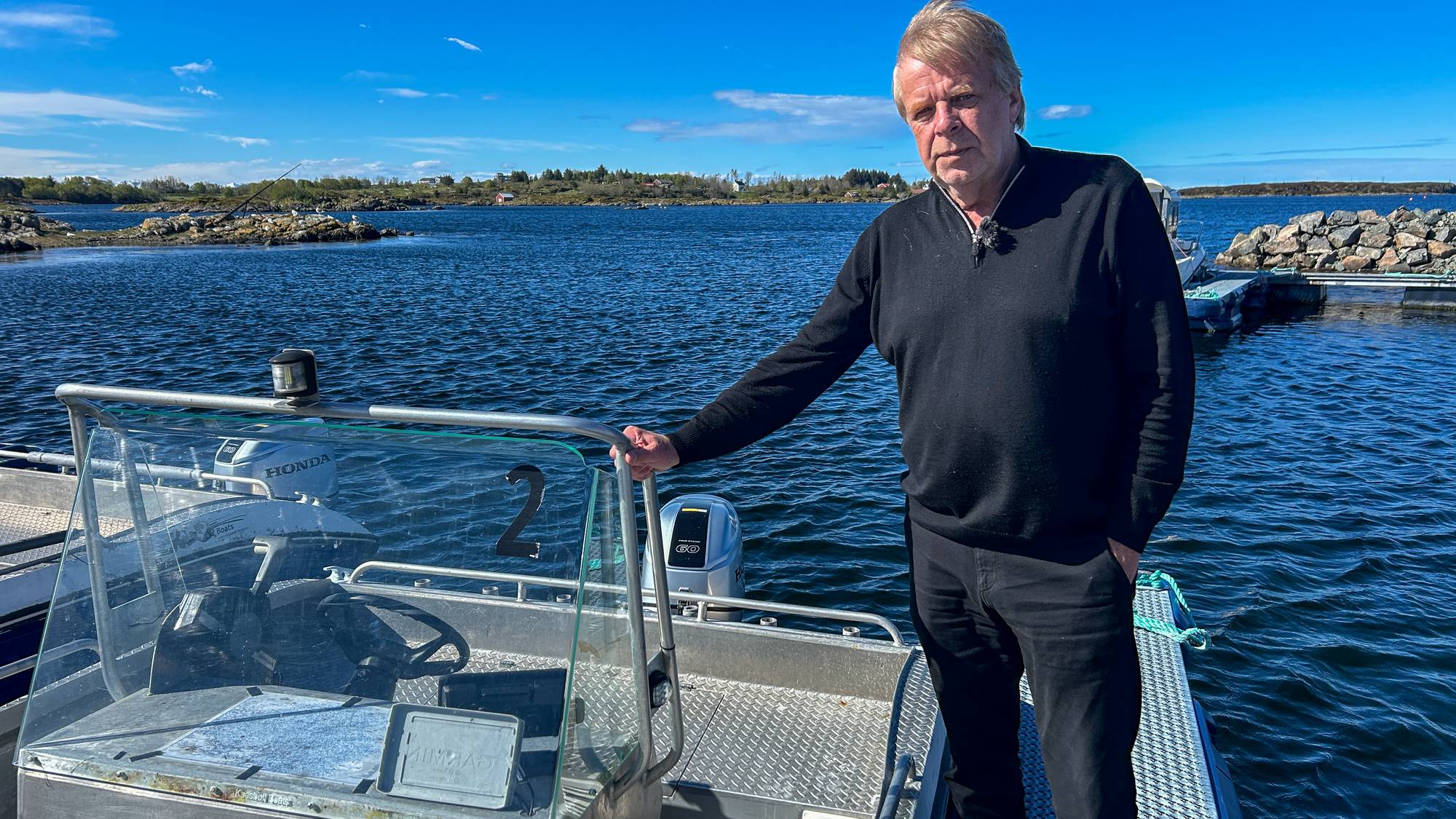 En eldre mann står på en båt. Bak ham er et rolig vann under en blå himmel. Han har på seg en svart genser og svarte bukser. Båten er ved en flytebrygge. (Bildebeskrivelsen er laget av en KI-tjeneste)
