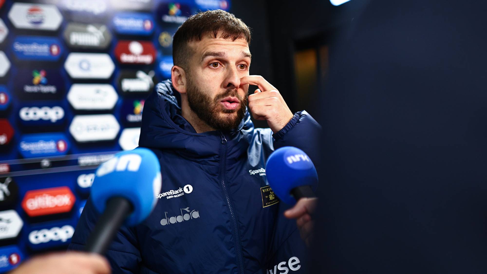Fotballtrener intervjues av journalister foran en reklamevegg etter kamp. (Bildebeskrivelsen er laget av en KI-tjeneste)