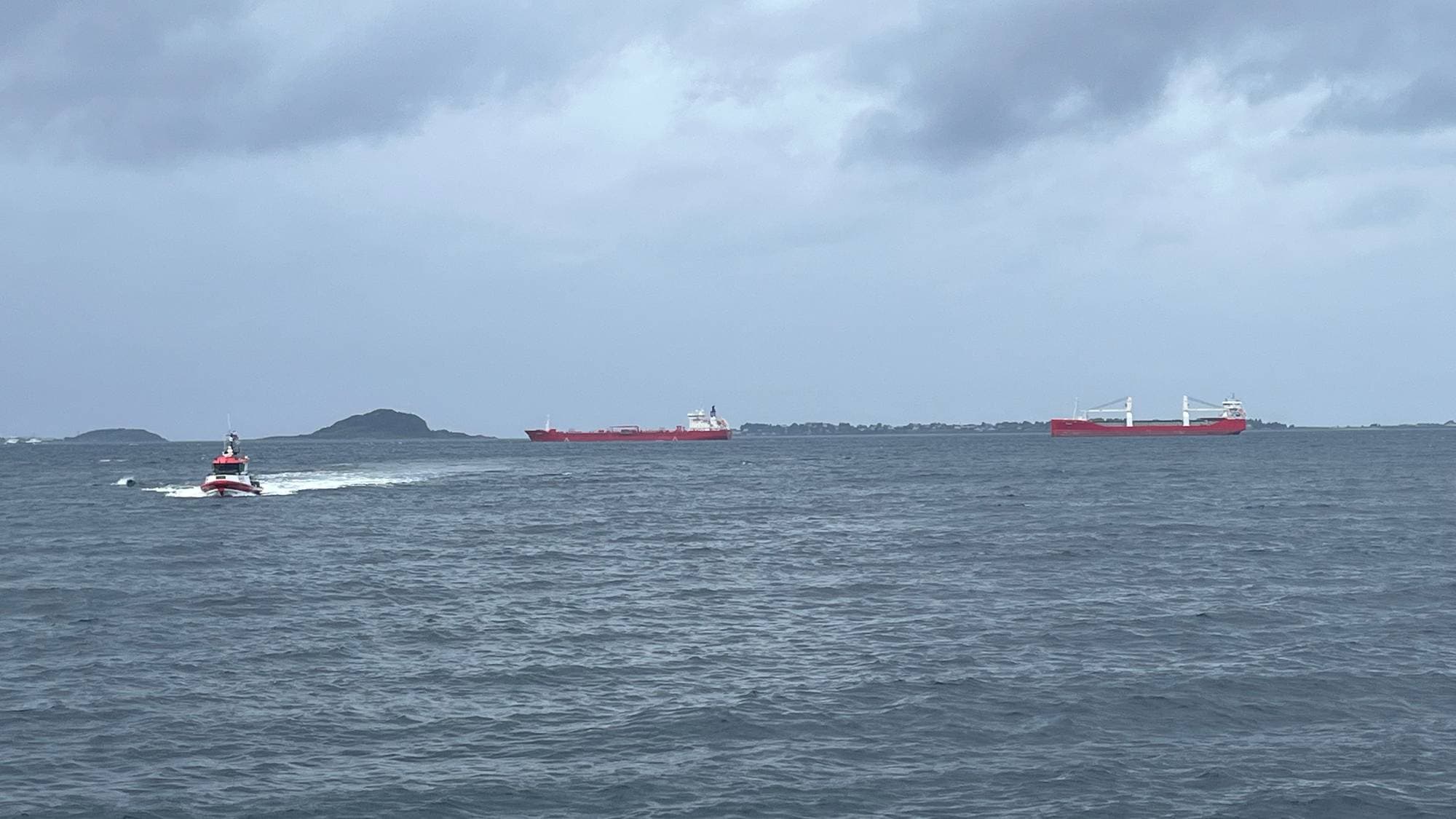 En losse/redningsbåt kjører på havet foran to store røde skip under en grå himmel. (Bildebeskrivelsen er laget av en KI-tjeneste)