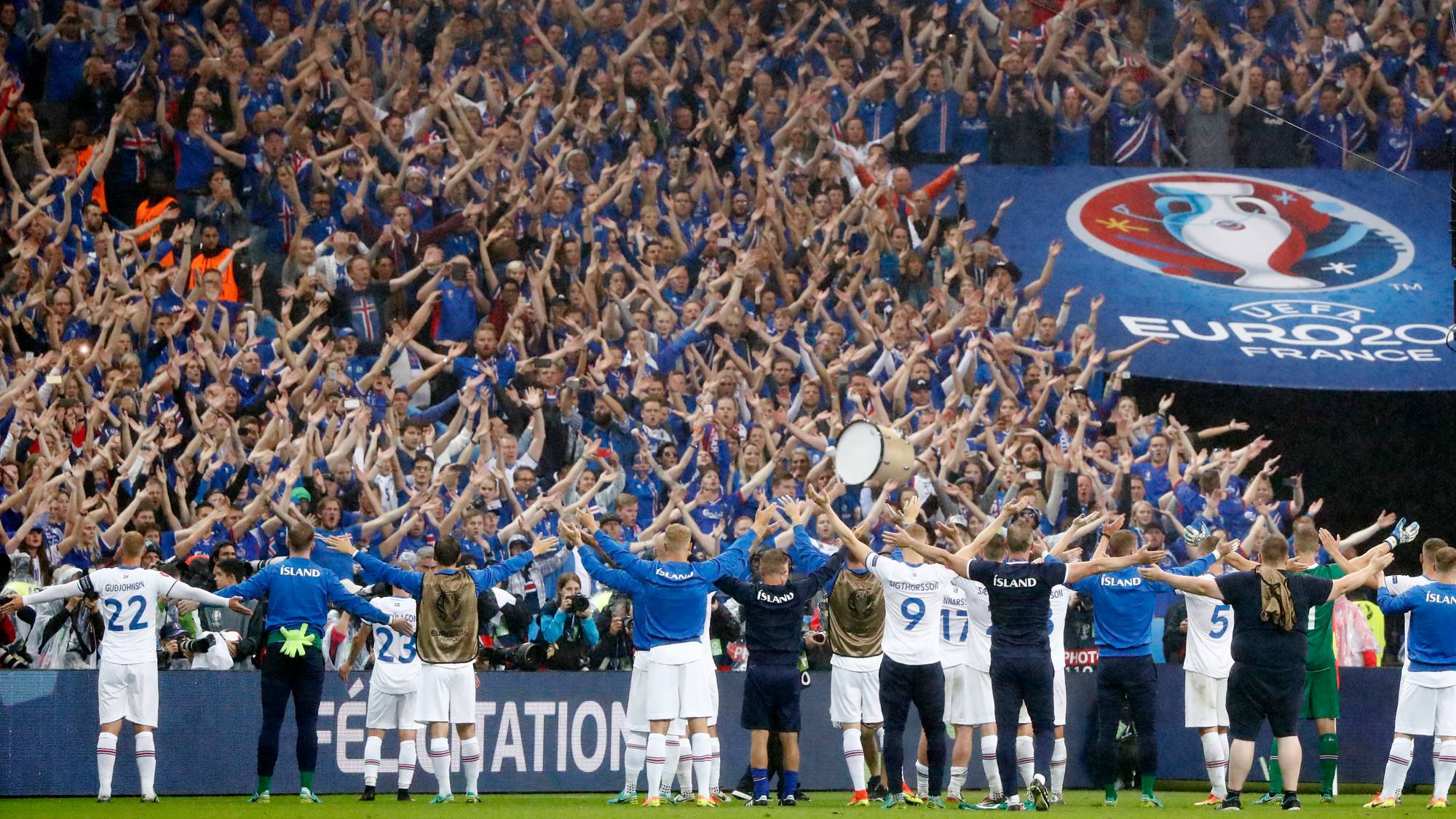 Islandsk fotballag feirer med supportere etter en kamp. Spillerne står på gresset med armene rundt hverandre, vendt mot en stor folkemengde. Supporterne, kledd i blått, står på tribunene med armene i været. Et banner for UEFA EURO 2020 vises i bakgrunnen. (Bildebeskrivelsen er laget av en KI-tjeneste)