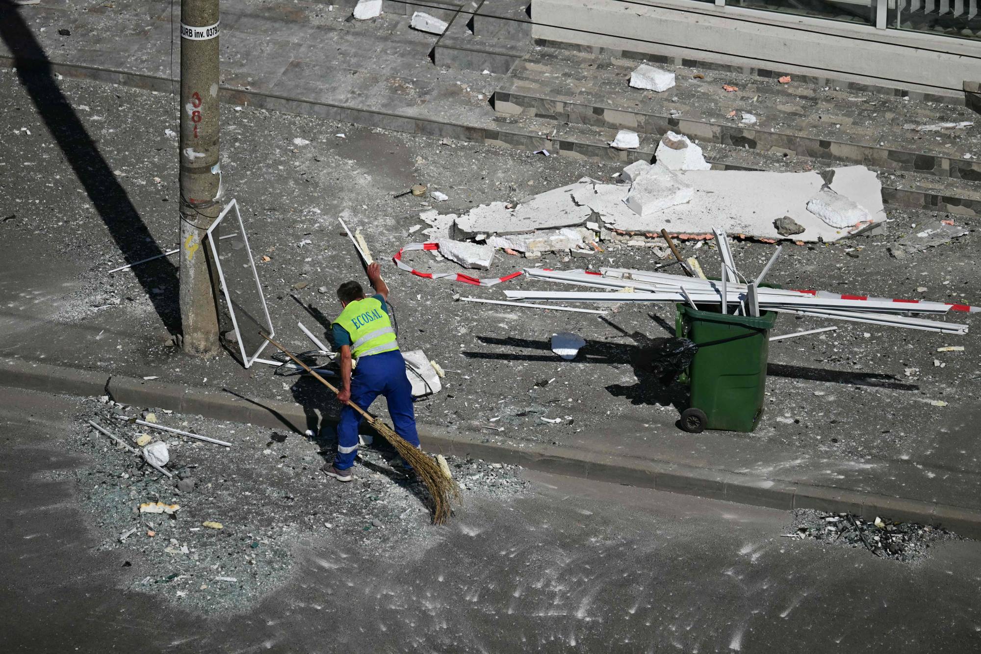 En person i gul vest feier gate med en kost. Der ligger mye bygningsrester og knust glass strødd utover et fortau og gate. En grønn søppeldunk er plassert til høyre for feieren. (Bildebeskrivelsen er laget av en KI-tjeneste)