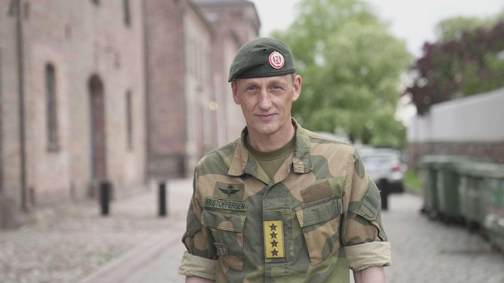 En mann ser inn i kamera. Murbygning og grønne trær i bakgrunn. 
