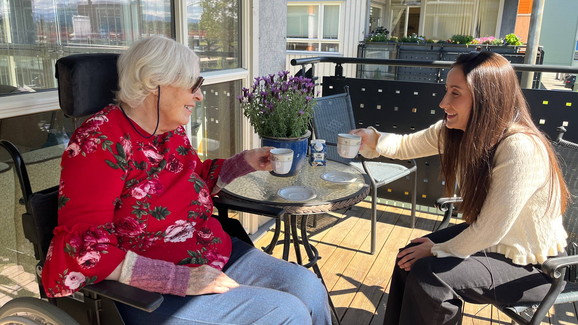 En eldre dame i rullestol med en rød blomstrete genser til venstre i bildet. Befinner seg på en balkong med en yngre kvinne med langt brunt hår og en hvit genser, som sitter til høyre for den eldre kvinnen. Mellom dem er et rundt bord som det står blomster og kaffefat på. De to kvinnene holder hver sin kaffekopp mens de skåler. 