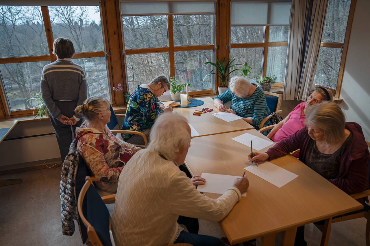 Syv beboere sitter sammen og tegner rundt bordet i oppholdsrommet. Den ene har sovnet, en annen stirrer rolig ut av vinduene med hendene knyttet bak ryggen.