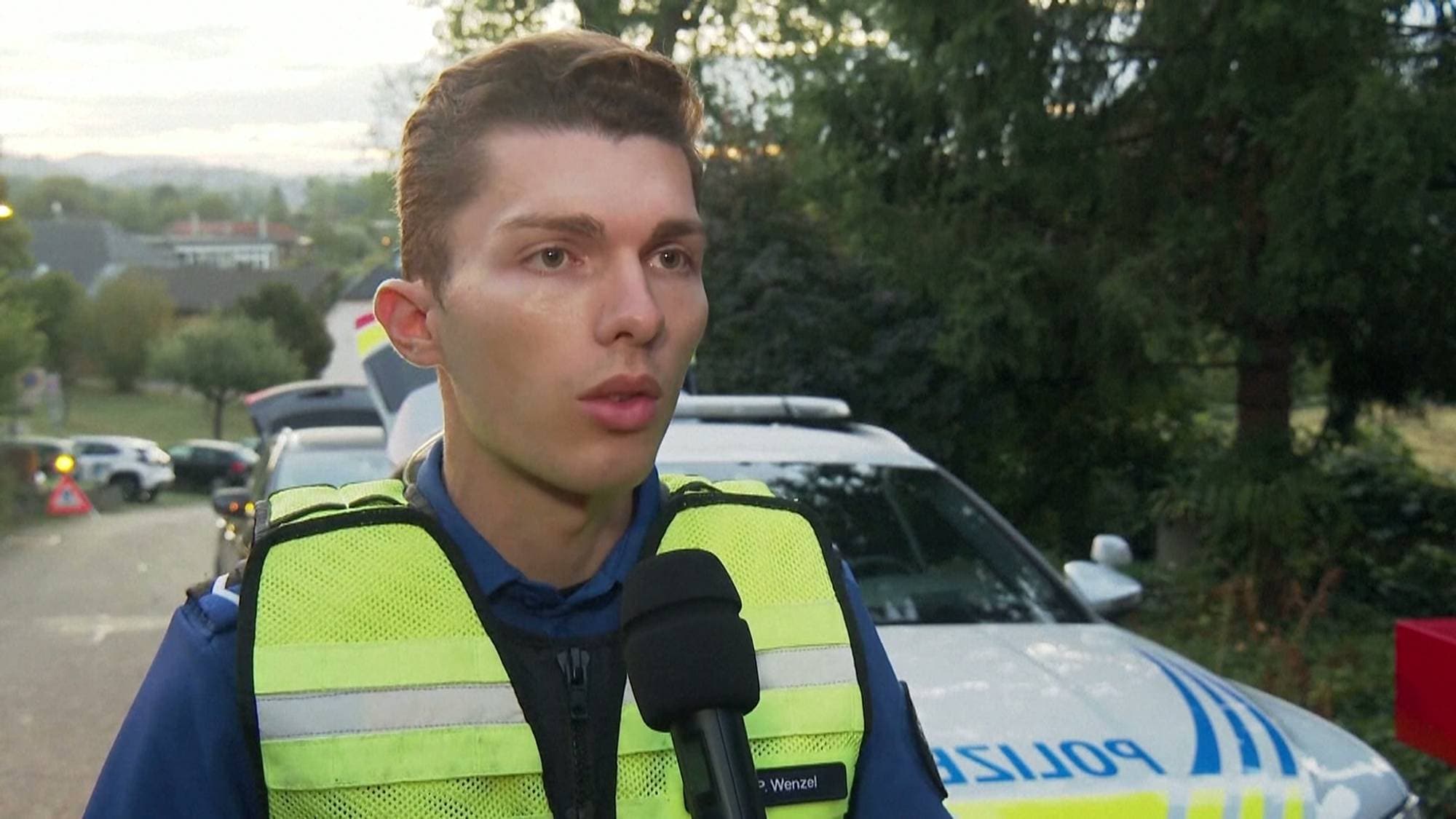 En politibetjent i gul refleksvest snakker i en mikrofon foran en politibil. (Bildebeskrivelsen er laget av en KI-tjeneste)