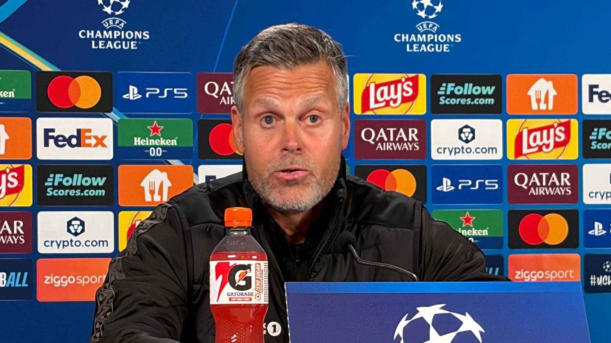 En mann sitter ved et bord foran en pressevegg med Champions League-logoer. (Bildebeskrivelsen er laget av en KI-tjeneste)