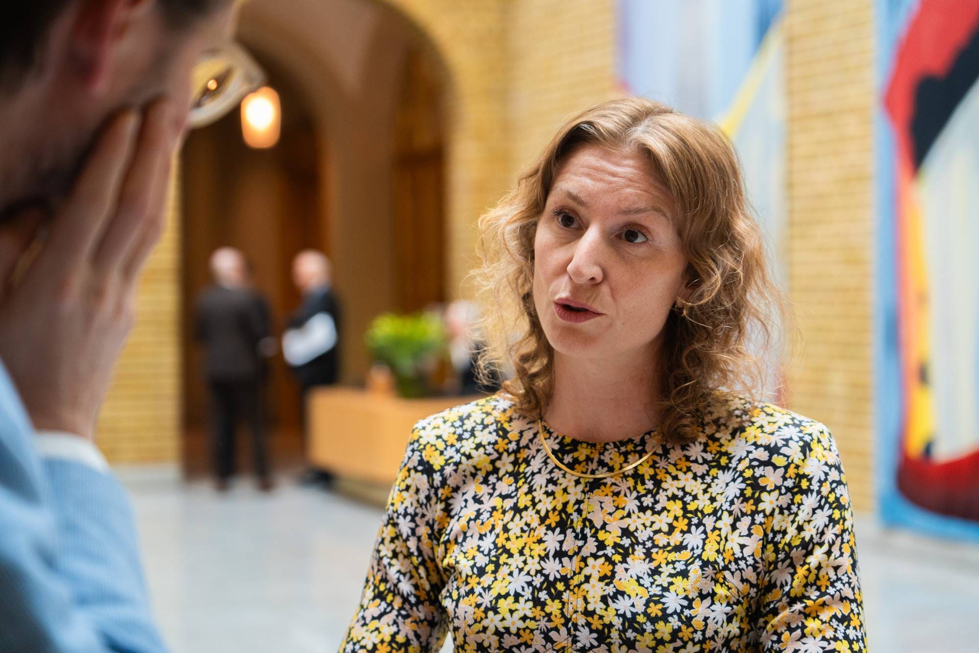 En kvinne med krøllete, lyst hår og en blomstrete kjole snakker. Hun har på seg et tynt, gullfarget halskjede. En mann, hvis ansikt er delvis skjult, står til venstre og ser ut til å lytte. I bakgrunnen står to menn som snakker sammen. Bildet er tatt innendørs i et rom med lyse vegger og en stor, fargerik abstrakt kunstinstallasjon. (Bildebeskrivelsen er laget av en KI-tjeneste)