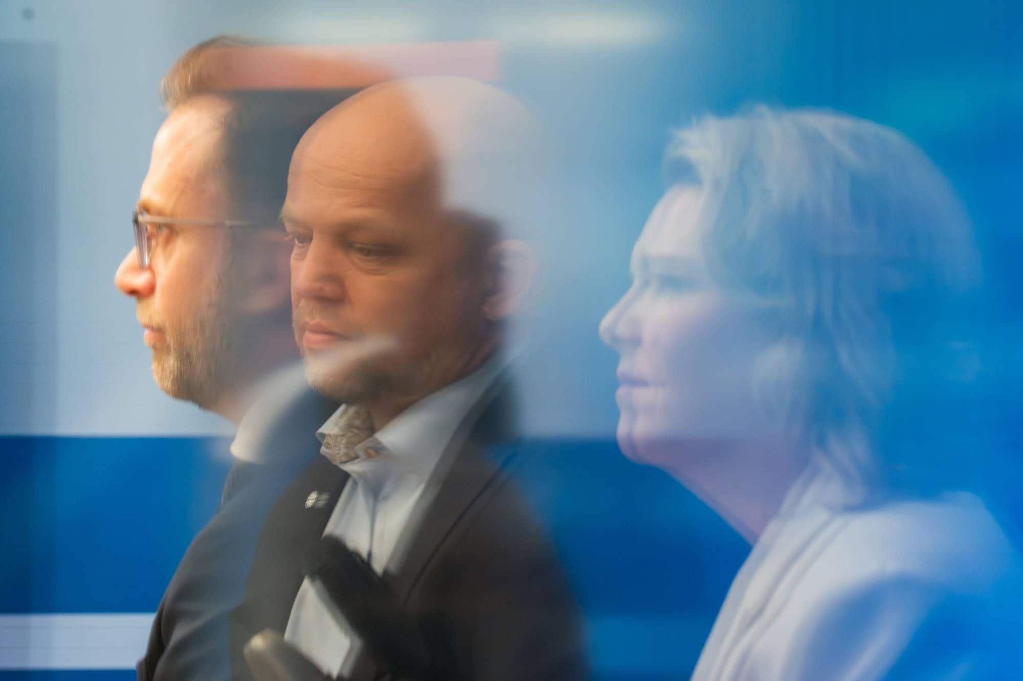 Tre personer ses i profil med effekten av bevegelse. En mann med briller til venstre, en mann med bart i midten, og en kvinne til høyre. De har på seg dress og lys skjorte. (Bildebeskrivelsen er laget av en KI-tjeneste)